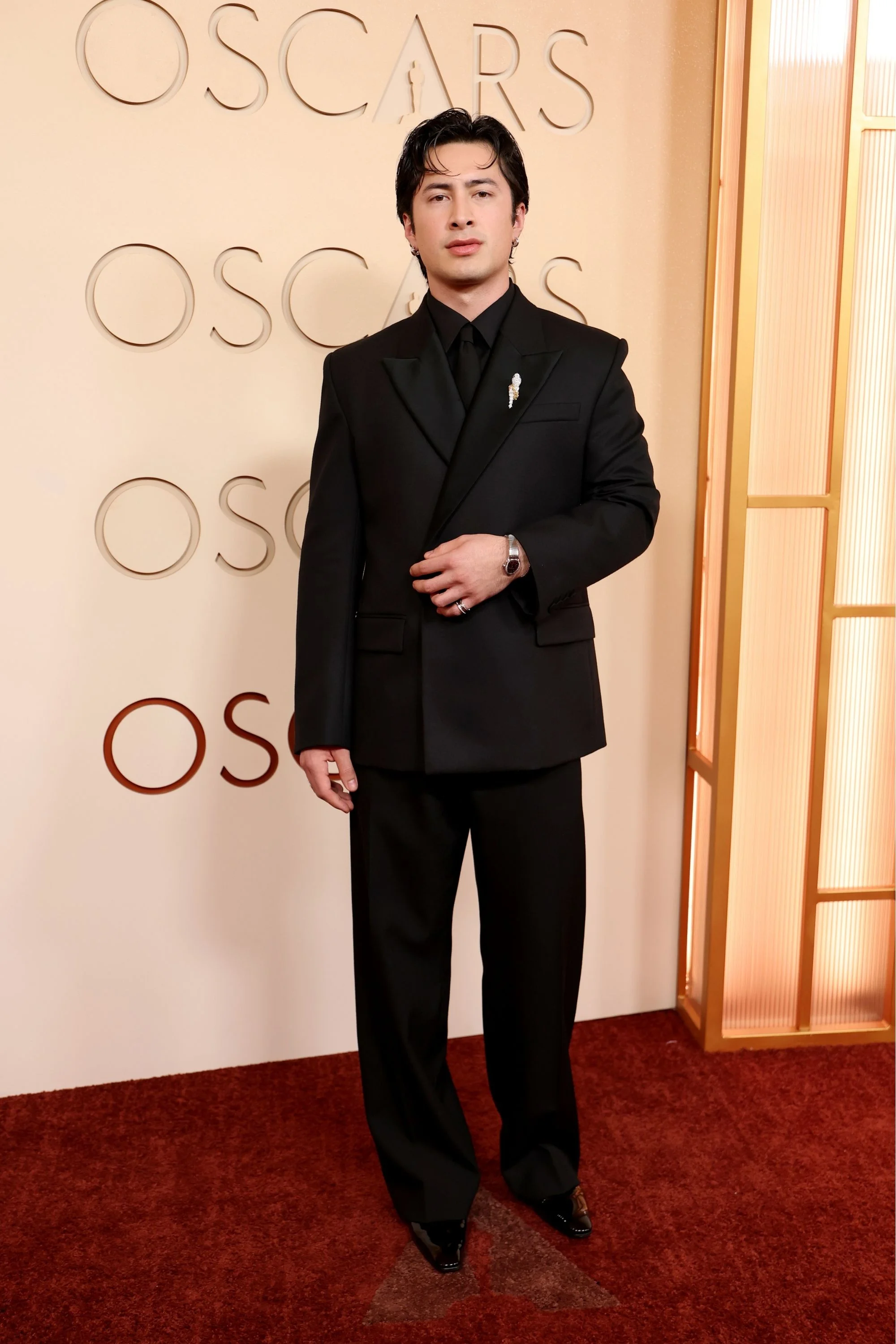 Oscar kırmızı halısında tam boy poz veren Hudson Williams, zarif siyah takım elbisesi, parlak rugan ayakkabıları ve sol kolundaki dikkat çekici Bulgari Serpenti kol saati ile modern ve şık bir görünüm sergiliyor.
