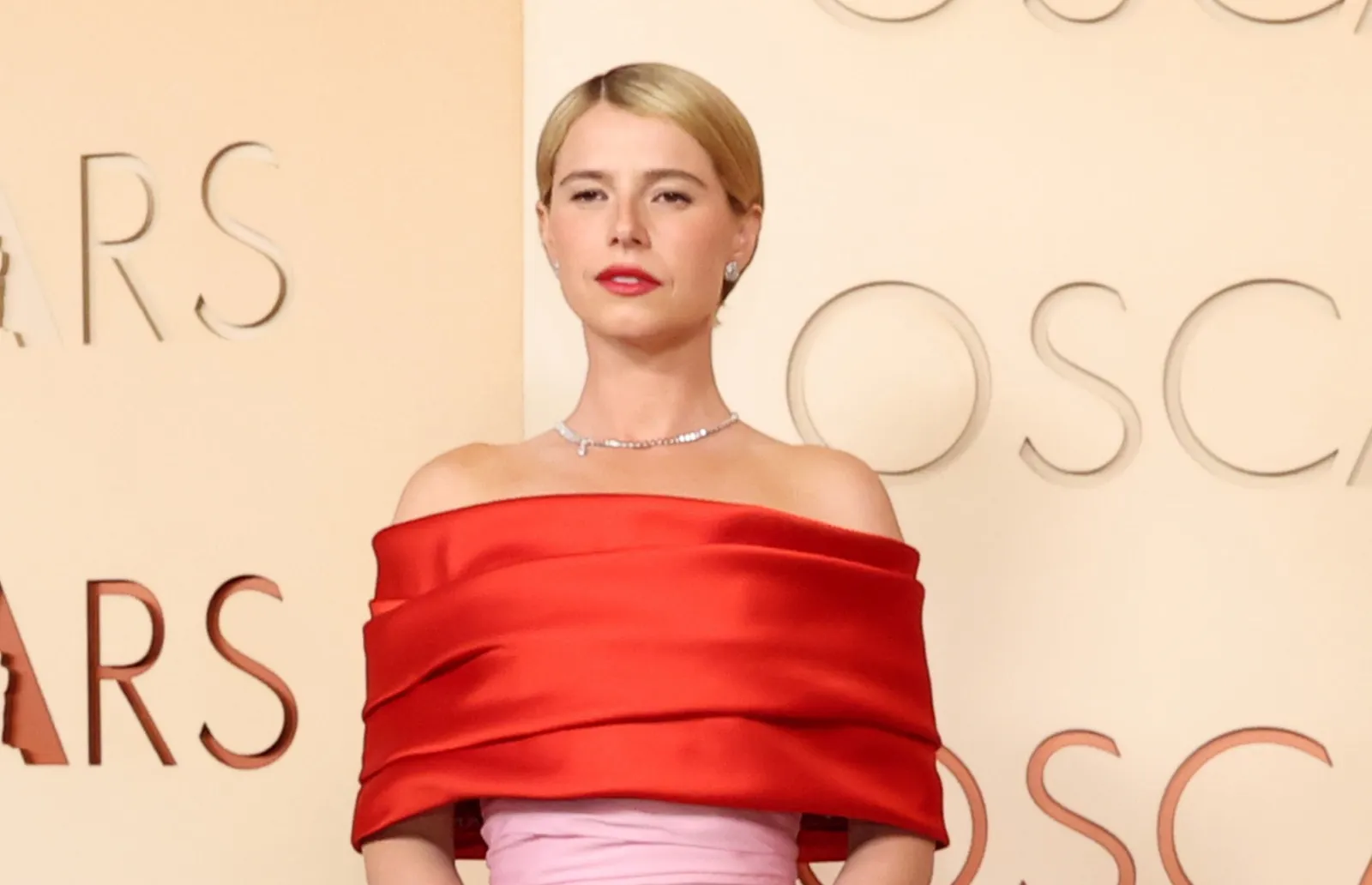 Oscar Ödülleri etkinliğinde poz veren Jessie Buckley, geriye taranmış sarı saçları, kırmızı ruju ve ciddi ifadesiyle dikkat çekiyor; omuzları açık parlak kırmızı elbisesini, ışıltılı gümüş bir kolye ve zarif küpelerle tamamlamış, arkasında ise kabartmalı OSCARS yazısının bir kısmı görülen bej bir duvar bulunuyor.