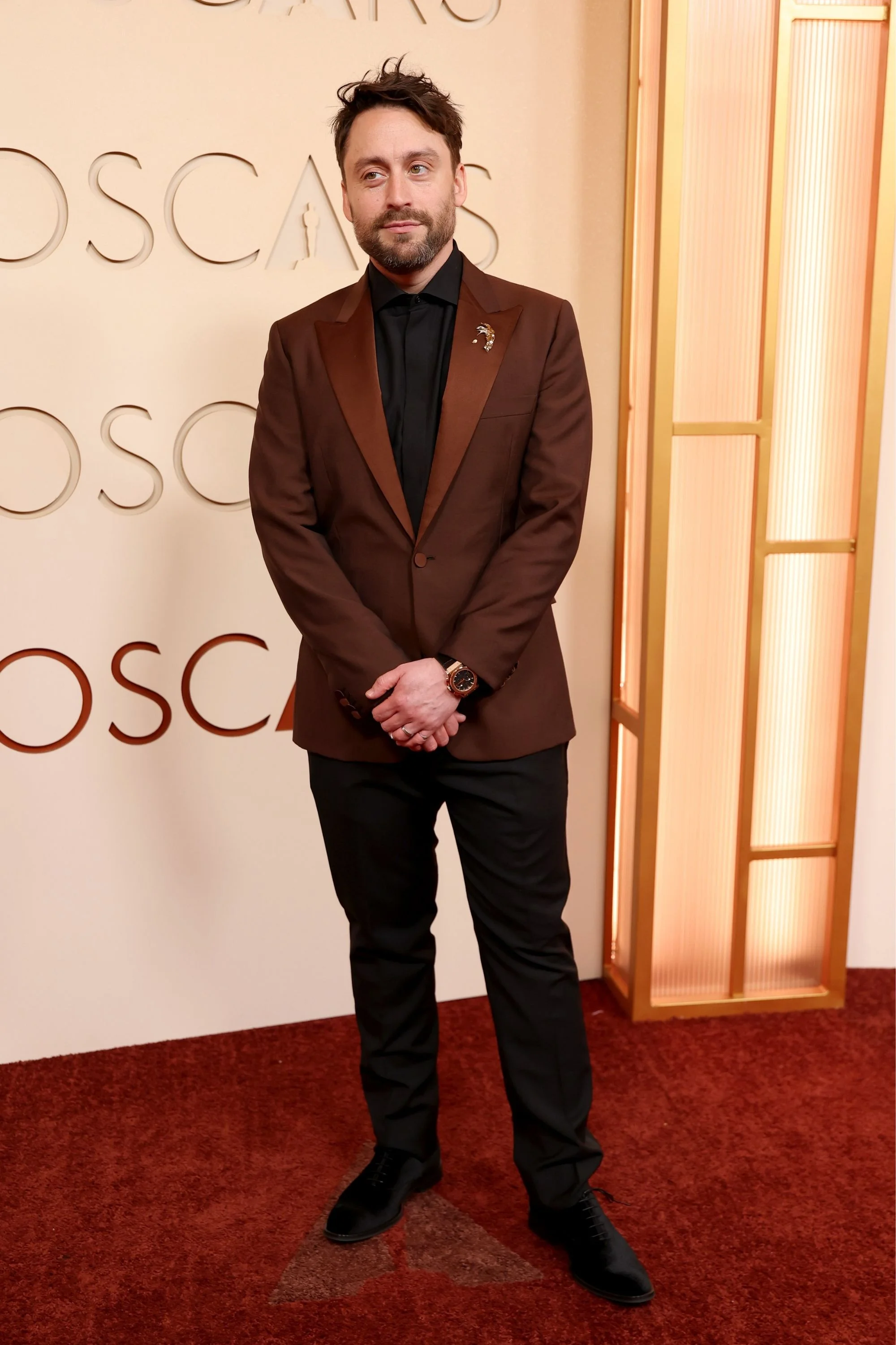Kieran Culkin, kırmızı halıda kahverengi bir takım elbise, siyah gömlek ve Hublot Classic Fusion Chronograph King Gold kol saatiyle zarif bir duruş sergiliyor; arkasında Oscar yazısı ve ışıklı bir panel belirginleşiyor.