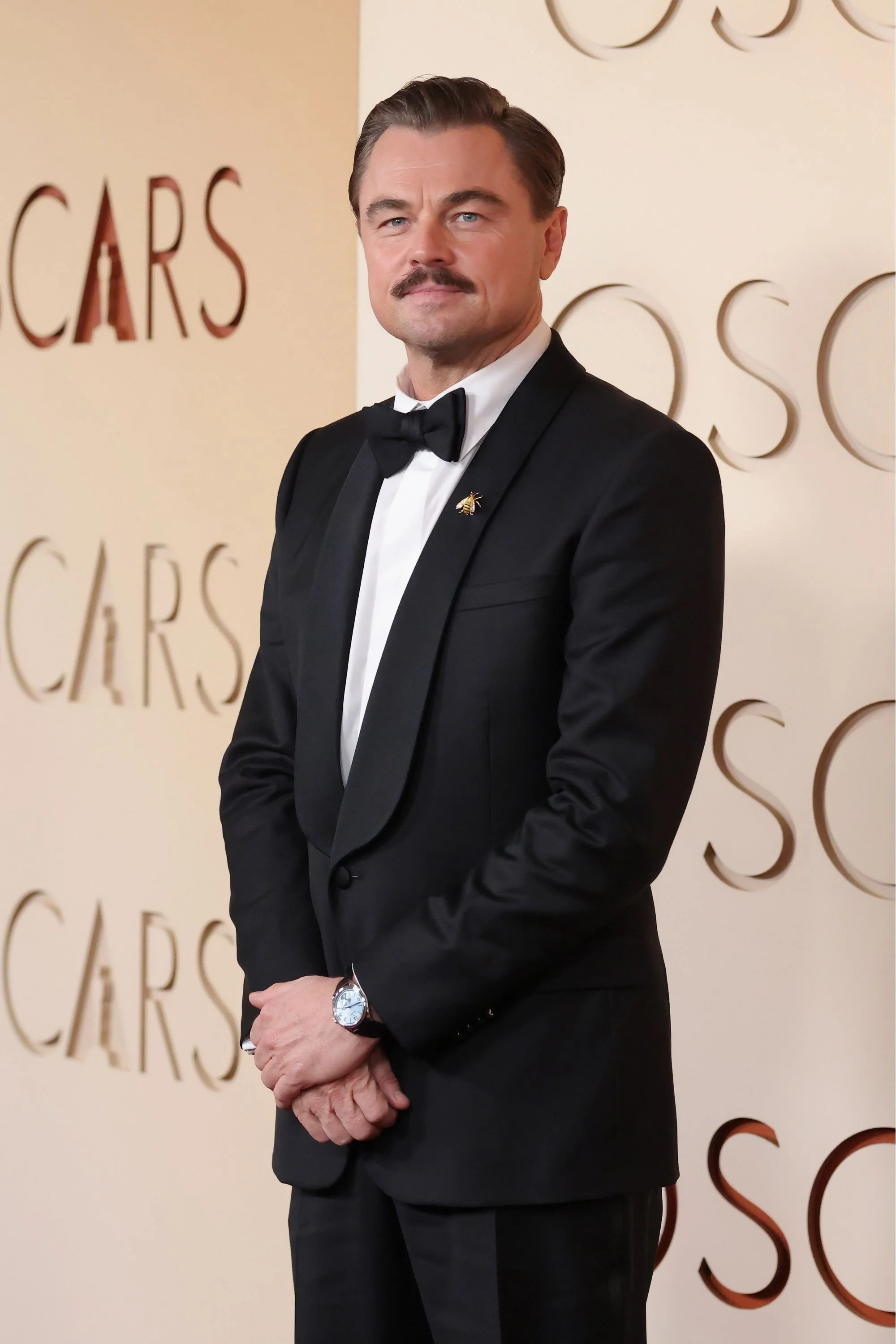 Oscar töreninde bıyıklı Leonardo DiCaprio, siyah smokini ve kravatı ile poz verirken sol bileğindeki dikkat çekici, zarif Rolex 1908 saati ve yakasındaki arı broşuyla şıklığını sergiliyor.