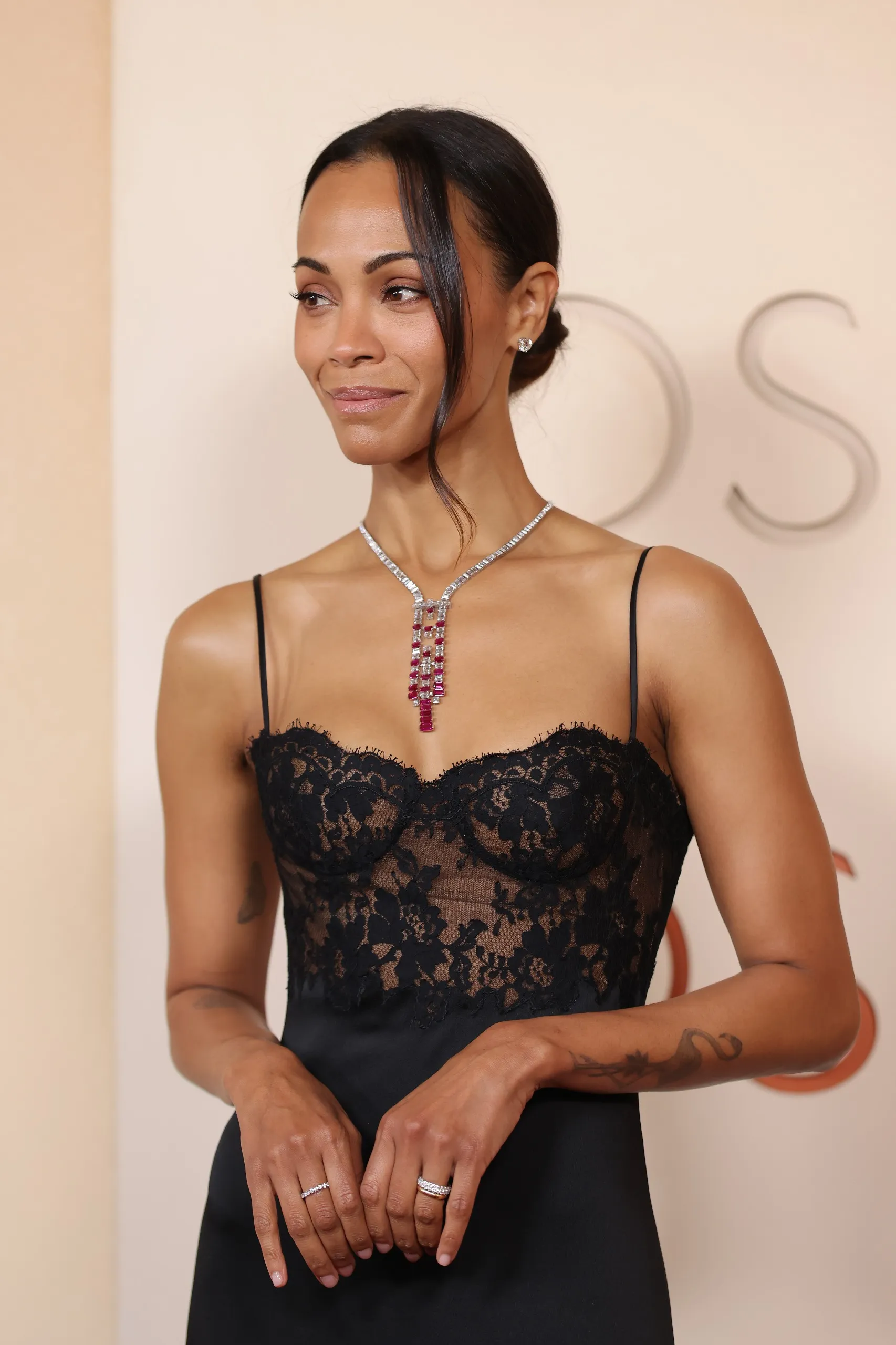 Oscar etkinliğinde siyah askılı dantel bir elbise giyen Zoe Saldana, boynunda gösterişli elmas ve yakut taşlarla bezeli bir kolye ile hafifçe gülümsüyor, parmaklarında yüzükler ve arkada belirgin OS harfleriyle dikkat çekiyor.