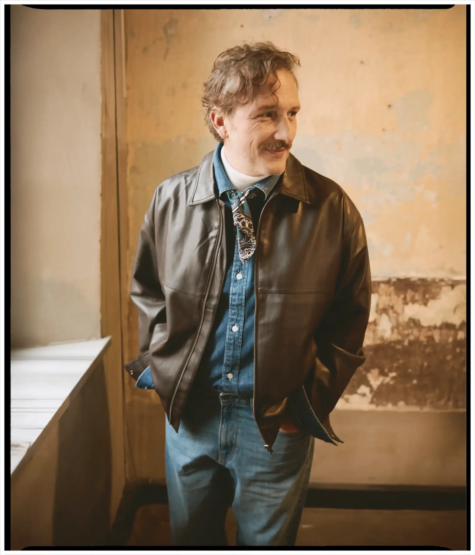 Osman Sonant röportaj çekiminde kahverengi deri ceket, denim gömlek ve jean kombinasyonu ile vintage dokulu mekânda sıcak tonlarda samimi bir stil portresi sunuyor.