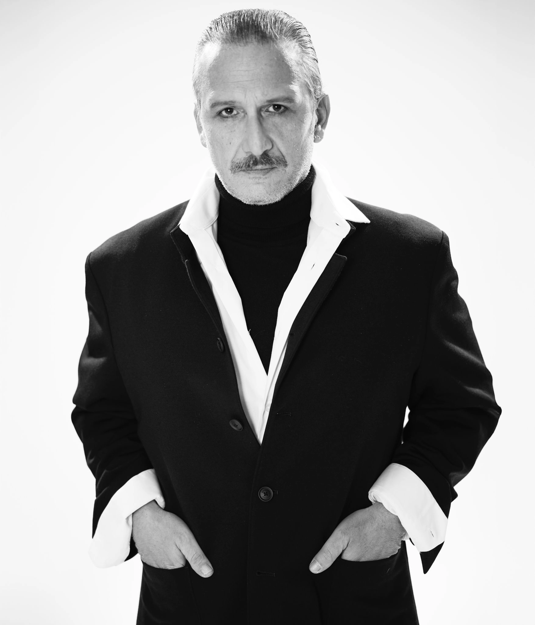 Osman Sonant siyah beyaz portrede siyah blazer, beyaz gömlek ve yüksek yakalı triko kombinasyonu ile minimal fonda güçlü ve zamansız bir stil duruşu sergiliyor.
