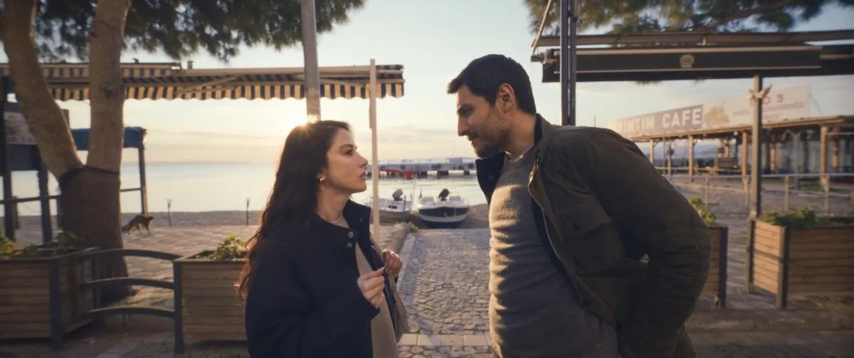 Özge Gürel ve Fatih Gühan'ın, güneş batarken veya doğarken deniz kenarındaki taş döşeli bir yolda karşılıklı durarak birbirlerine baktığı, arka planda tekneler, şemsiyeli kafeler ve uzakta yürüyen küçük bir köpeğin göründüğü bir film karesi.