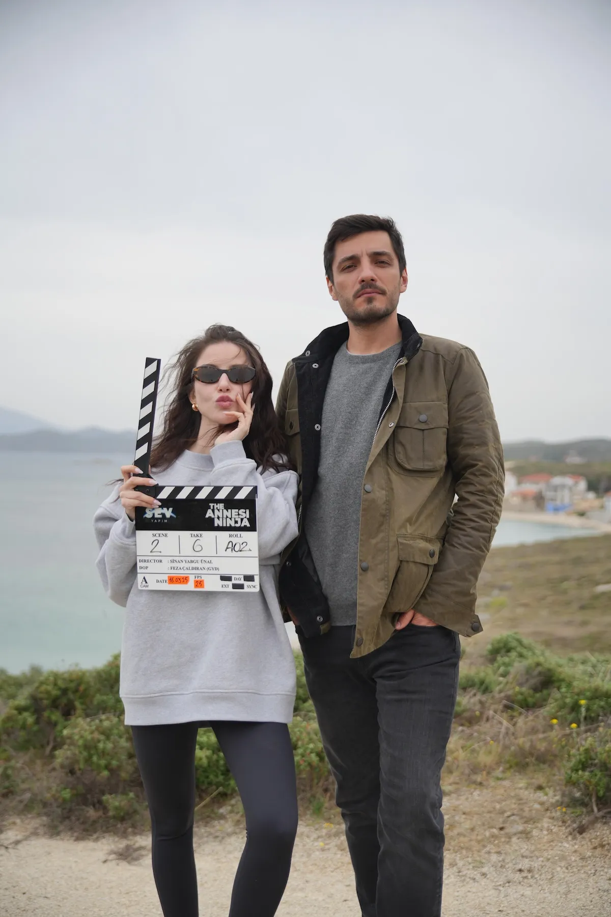 Özge Gürel ve Fatih Gühan, The Annesi Ninja filminin çekimleri sırasında, Gürel'in elinde projenin adının yazılı olduğu bir klaketle, sahil kenarında bulutlu bir havada objektife poz veriyor; Gürel dudaklarını büzmüş, Gühan ise ciddi bir ifadeyle duruyor.