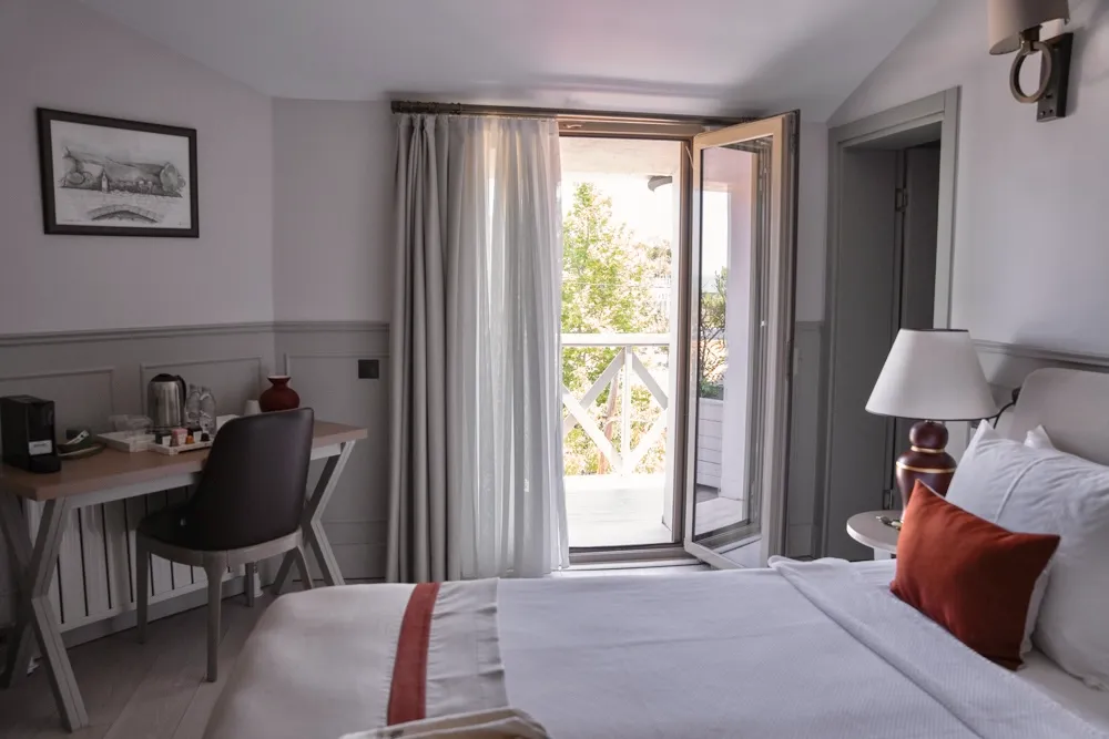Şile'deki Parma Sole otel odasında, gün ışığının içeri aktığı açık balkondan yeşilliklerin göründüğü, beyaz nevresimli yatak, turuncu vurgulu yastık, çalışma masası ve rahat bir sandalye ile aydınlık ve davetkar bir atmosfer.