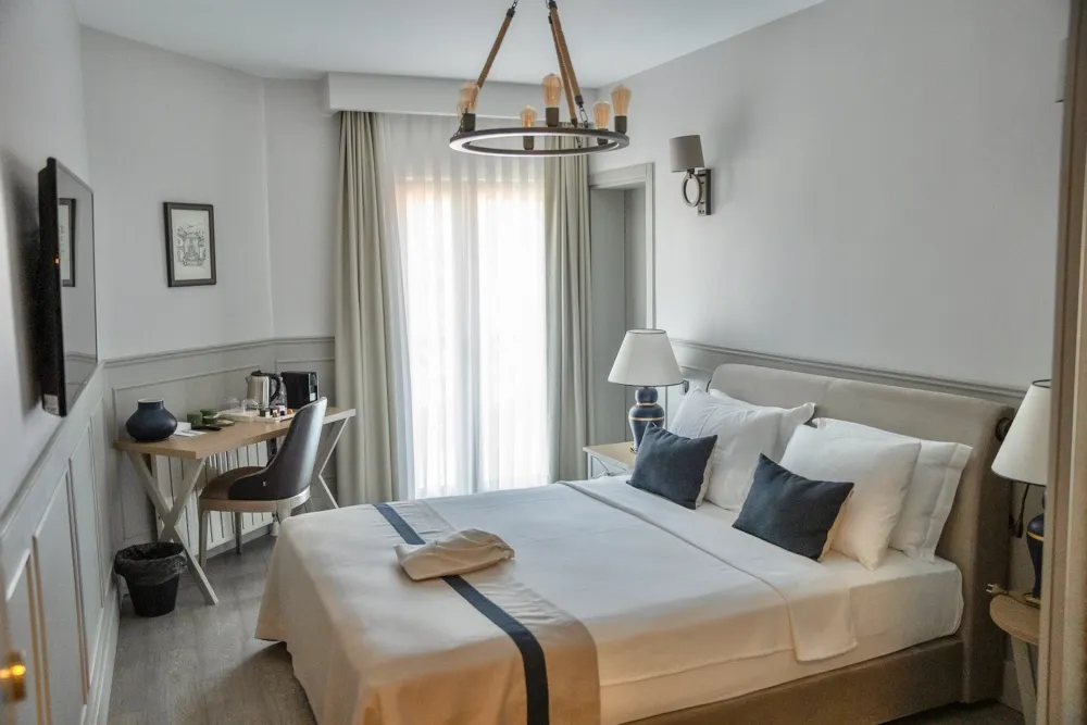 Şile'deki konaklama deneyimini vurgulayan bu modern otel odasında, beyaz nevresimli yatak koyu lacivert yastıklar ve bej renkli bir bornozla tamamlanırken, sol taraftaki çalışma masası, duvara monte televizyon ve şık avizeyle aydınlık pencere detayı dikkat çekiyor.