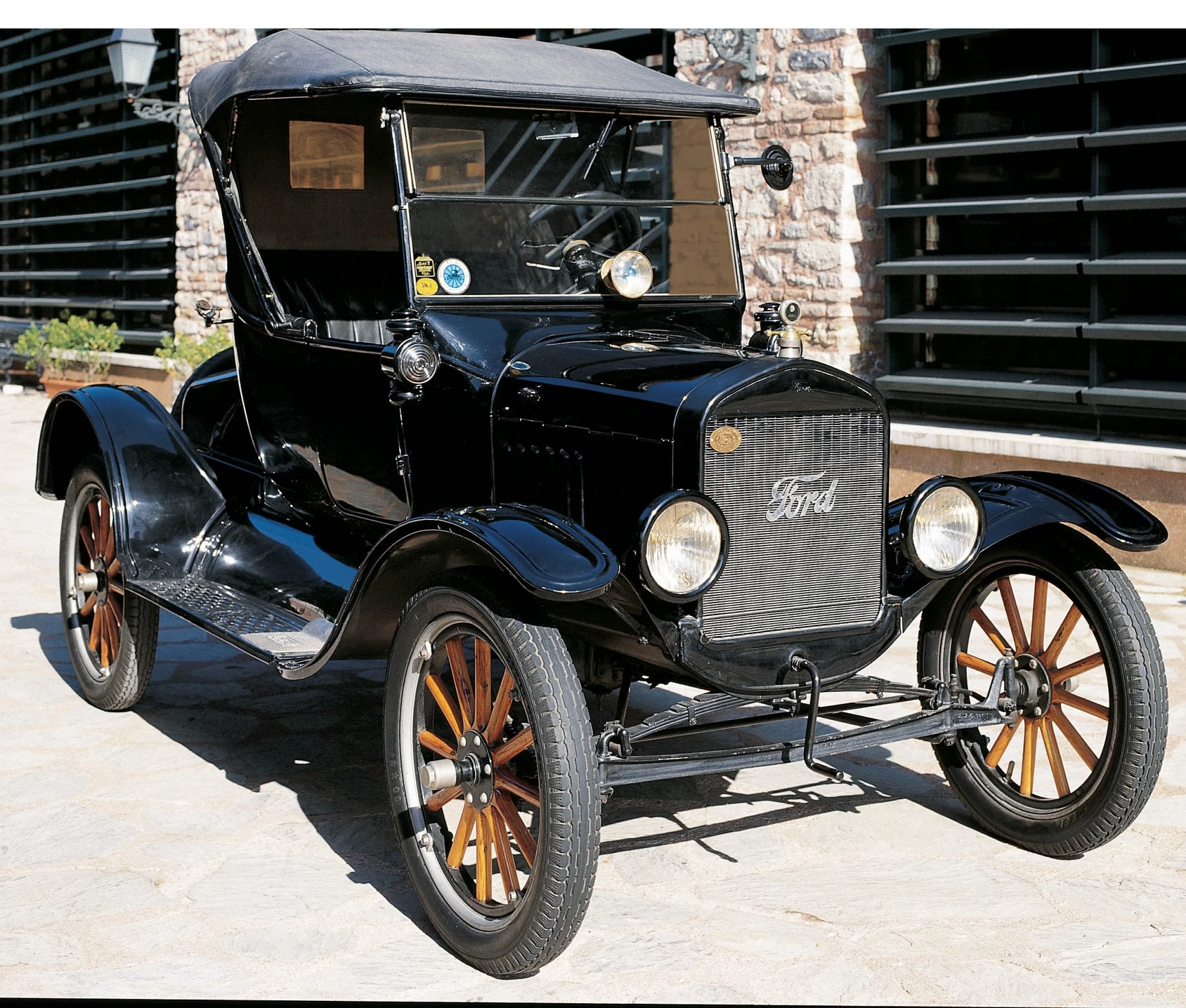 Peaky Blinders temasıyla uyumlu, parlak siyah boyası, ahşap telli tekerlekleri ve katlanır tavanıyla öne çıkan bu ikonik Ford Model T, taş duvarlı ve modern panjurlu bir yapının önünde, güneşli bir günde park edilmiş olarak sergileniyor.