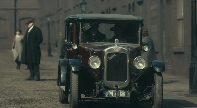 Peaky Blinders dizisinden bir kesit: Tozlu bir sokakta, ön planda koyu bordo renkte, parlak ön ızgarası ve yuvarlak farlarıyla dikkat çeken klasik bir Austin otomobil, arkasında ise dönemin şık kıyafetlerini giymiş belirsiz insan silüetleri.