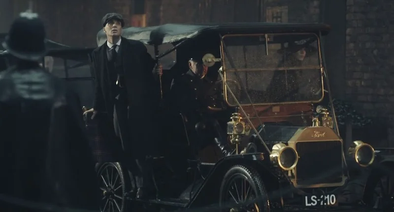 Peaky Blinders dizisinin ana karakteri Thomas Shelby, karanlık ve yağmurlu bir sokakta duran, detaylı altın rengi farlara sahip Ford Model T model aracından öne doğru hafifçe eğilmiş, dikkatle etrafa bakarken görülüyor.