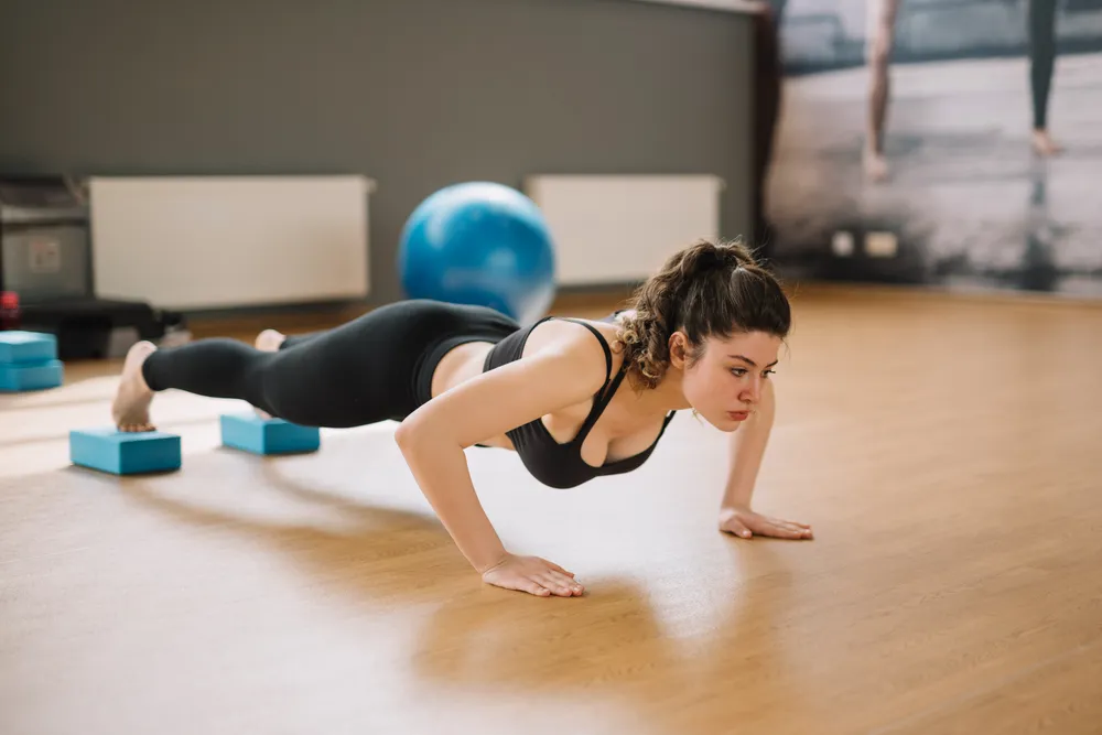 Koyu renk spor kıyafetleri giymiş genç bir kadın, fitness salonunda elleri zeminde, ayakları ise mavi egzersiz blokları üzerinde yükseltilmiş şekilde, vücudunu düz tutarak ve odaklanmış bir ifadeyle zorlu bir plank duruşu egzersizi yapıyor.