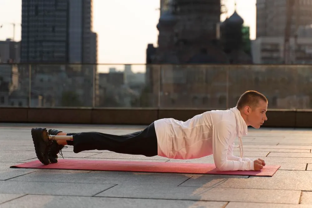 Şehir silüetinin göründüğü bir terasta, kırmızı bir yoga matının üzerinde, protez bacaklı kısa saçlı genç bir birey, kolları ve ayak parmakları üzerinde güçlü bir şekilde durarak doğru formda bir plank duruşu sergiliyor.