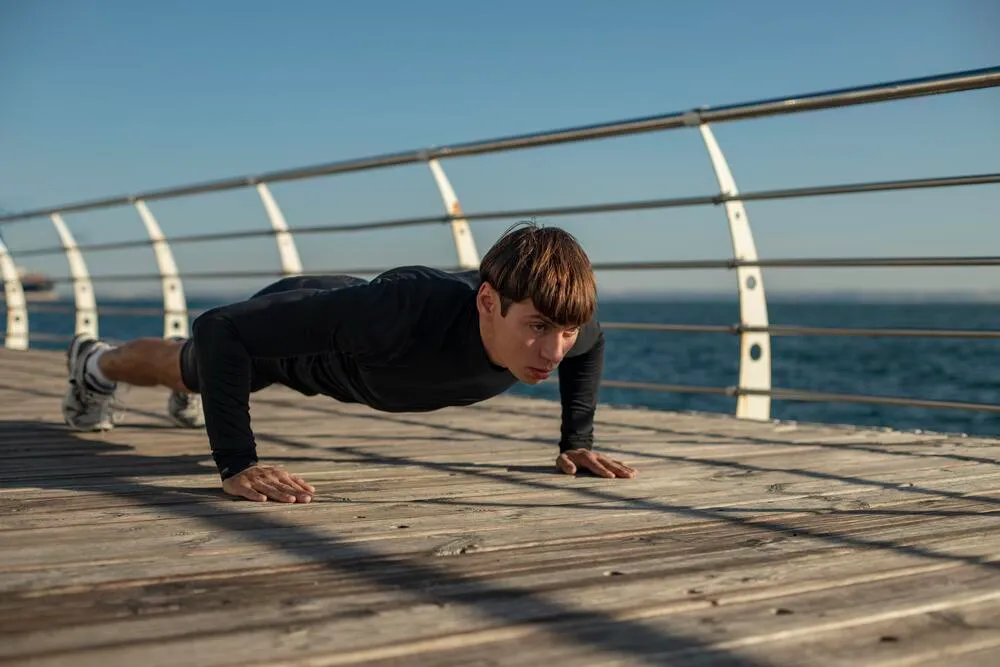 Genç bir adam, güneşli havada deniz kenarındaki ahşap bir platform üzerinde, siyah uzun kollu tişört ve spor ayakkabılarıyla doğru formda bir plank duruşu yapıyor, arka planda masmavi deniz ve açık gökyüzü uzanıyor.