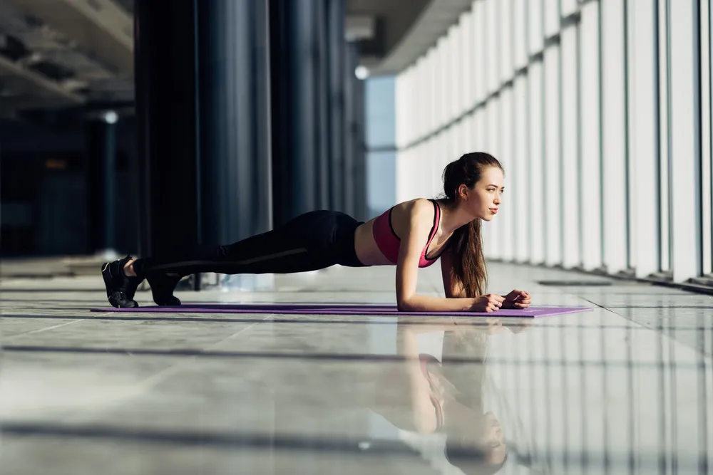 Genç bir kadın, modern ve aydınlık bir kapalı alanda, parlak zemine serilmiş mor yoga matı üzerinde, pembe spor sütyeni ve siyah taytlarıyla dirsekleri ve ayak parmakları üzerinde doğru plank duruşunu sergiliyor, yüzünde odaklanmış bir ifade ve zemin yansımalarıyla dikkat çekiyor.
