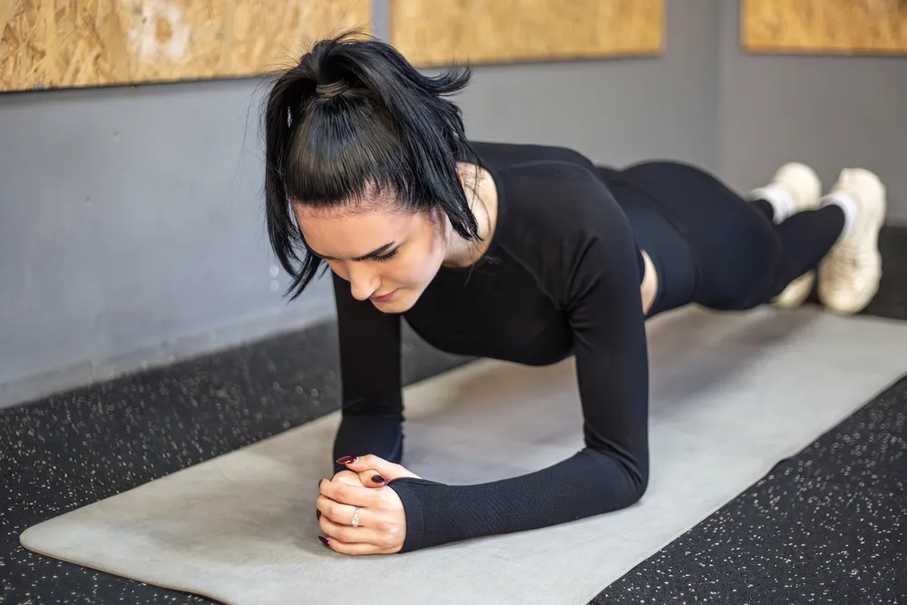 Başını hafifçe aşağı eğmiş, siyah uzun kollu spor kıyafeti giyen genç bir kadın, koyu renk benekli zeminde duran açık gri yoga matının üzerinde kollarını ve ayak parmaklarını kullanarak kusursuz bir plank duruşu sergiliyor.