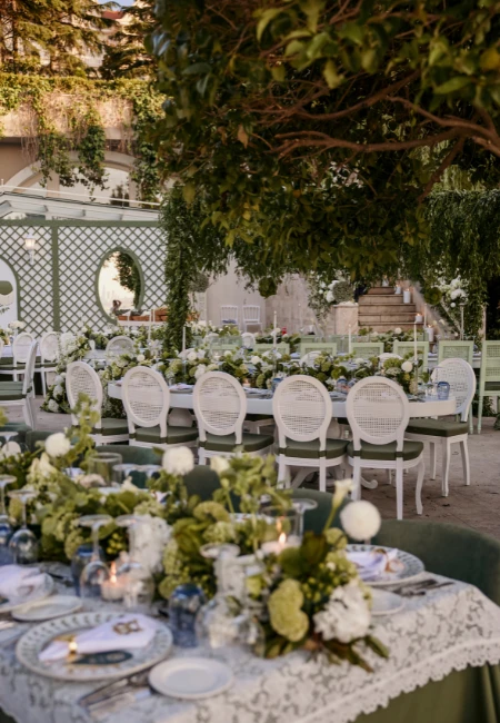 Büyükada'nın yeşillikler içindeki Princes' Palace Resort'ta, beyaz dantel örtülü uzun masaların yeşil kumaşlarla birleştiği, zarif beyaz sandalyeler ve yoğun çiçek aranjmanlarıyla süslenmiş, deneyim odaklı bir açık hava düğün yemeği alanı görülüyor.