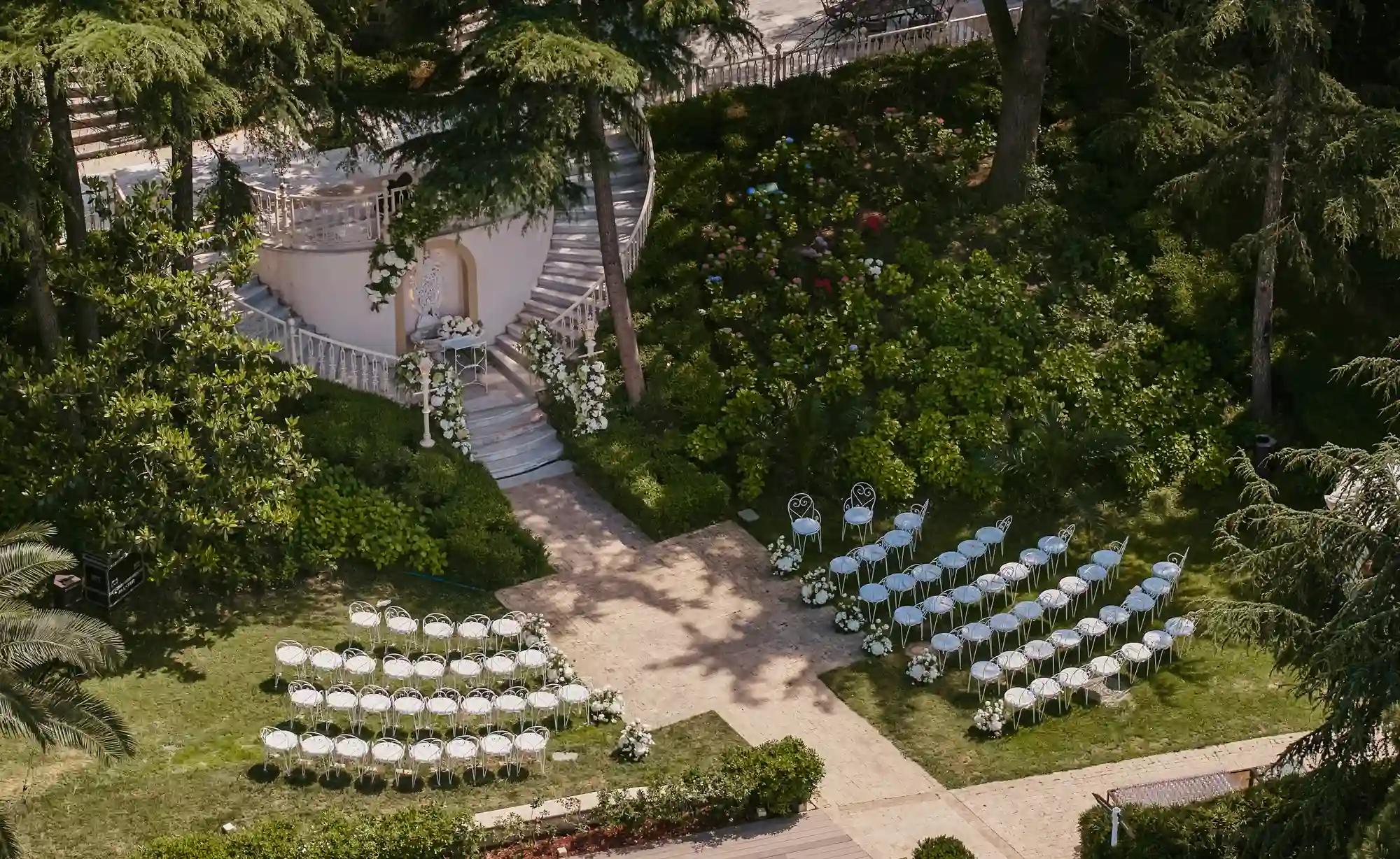 Büyükada’da doğanın kalbinde yer alan Princes’ Palace Resort'un yemyeşil bahçesinde, beyaz sandalyelerin özenle sıralandığı ve çiçeklerle süslenmiş zarif bir nikah alanı ile deneyim odaklı bir düğün için hazırlanan açık hava tören alanı kuşbakışı görünüyor.