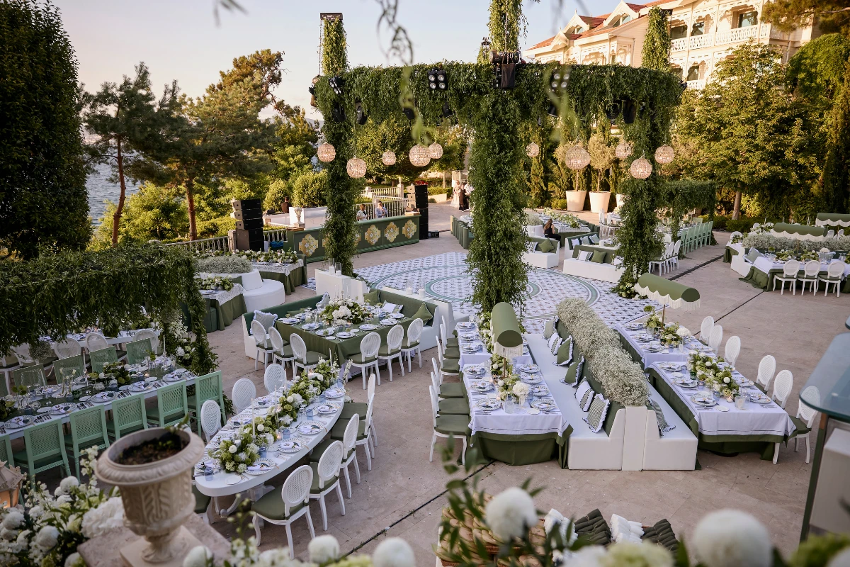 Büyükada'daki Princes' Palace Resort'ta, yemyeşil bitkilerle süslenmiş büyük bir kemerin çevrelediği desenli zemin üzerinde, beyaz ve yeşil detaylarla bezenmiş, zarif masa düzenlemeleri ve dinlenme alanlarıyla hazırlanmış, deniz manzaralı deneyim odaklı bir düğün mekanı gün batımı ışıkları altında.