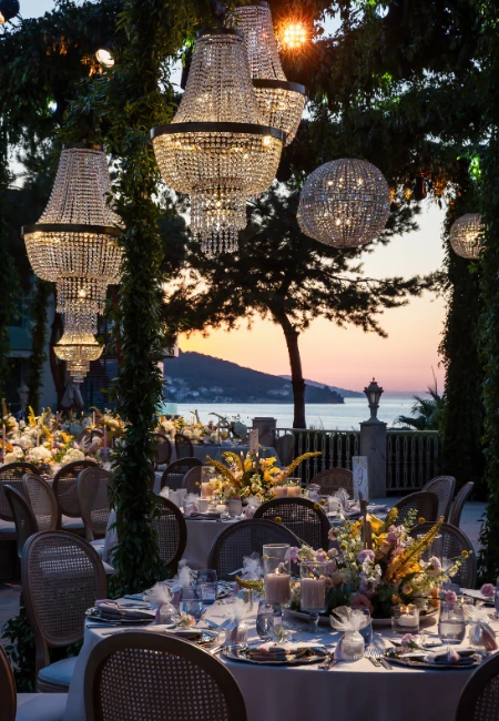 Akşamüstünün yumuşak ışıkları altında, Princes' Palace Resort'un Büyükada'da denize nazır açık hava terasında, kristal avizeler ve yeşil sarmaşıklarla bezeli çardak altında zarifçe hazırlanmış yuvarlak düğün masaları, adanın romantik gün batımı manzarası eşliğinde davetlileri bekliyor, bu an deneyim odaklı bir kutlamanın başlangıcını işaret ediyor.