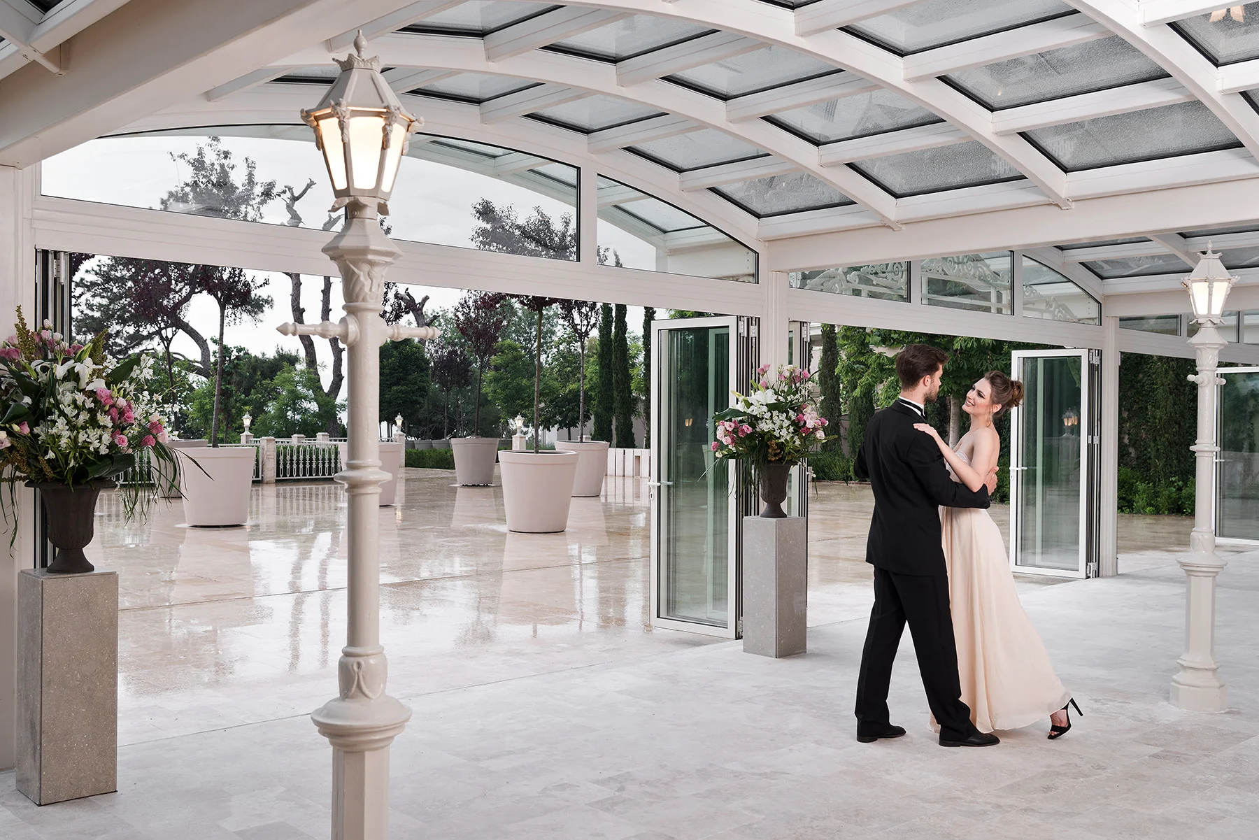 Büyükada'da bir düğün için Princes' Palace Resort'un cam tavanlı, ferah balo salonu veya teras alanında, bej elbisesi ve siyah takımıyla dans eden bir çift, etrafı çiçek aranjmanları ve aydınlatmalı direklerle süslü yeşil bir bahçeye açılan deneyim odaklı bir mekânın keyfini çıkarıyor.