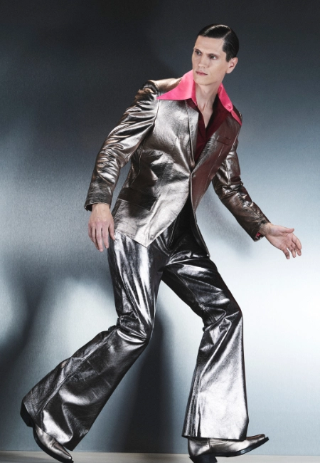 Tam boy, parlak gümüş rengi metalik ceket ve ispanyol paça pantolon giyen, pembe yakalı bir gömlekle stilize edilmiş bir erkek model, Hatice Gökçe'nin Remnant koleksiyonundaki erkekliğin dönüşümünü ve parçalanmış ama güçlü varlığını yansıtan dikkat çekici bir yüzey sunuyor.