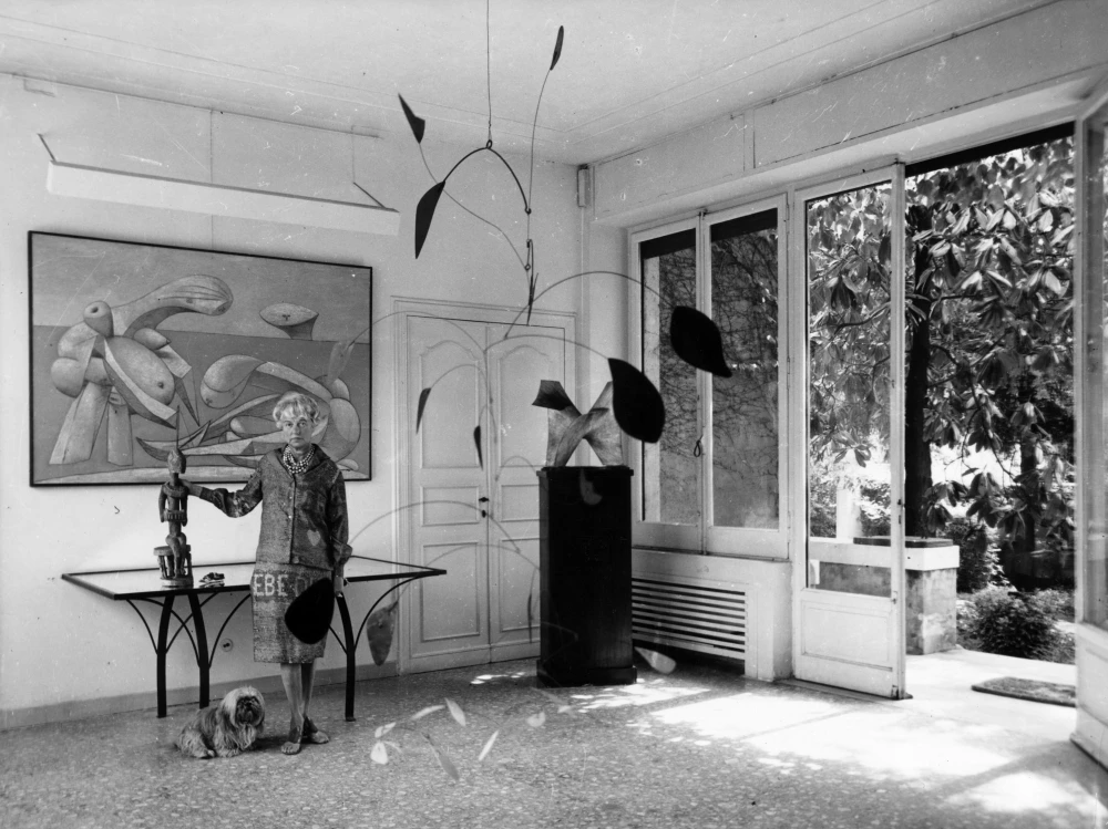 Sanat koleksiyoncusu Peggy Guggenheim, inci kolyesi ve desenli elbisesiyle, evindeki zengin sanat koleksiyonunu sergilerken, bir eliyle ahşap bir heykeli tutmuş, önünde sevimli köpeği uzanırken, arkasında büyük bir soyut tablo ve havada süzülen hareketli bir heykelin önünde poz veriyor.