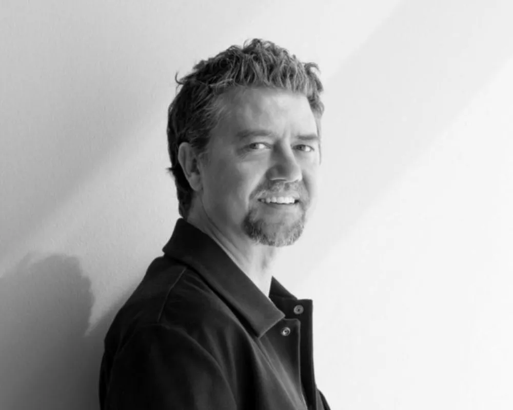 Sanatçı Seçkin Pirim'in, kameraya doğru hafifçe gülümseyerek bakan, dağınık kıvırcık saçları ve keçi sakalıyla sade bir arka plan önünde durduğu, koyu renk yakalı bir ceket giydiği siyah beyaz portre fotoğrafı.