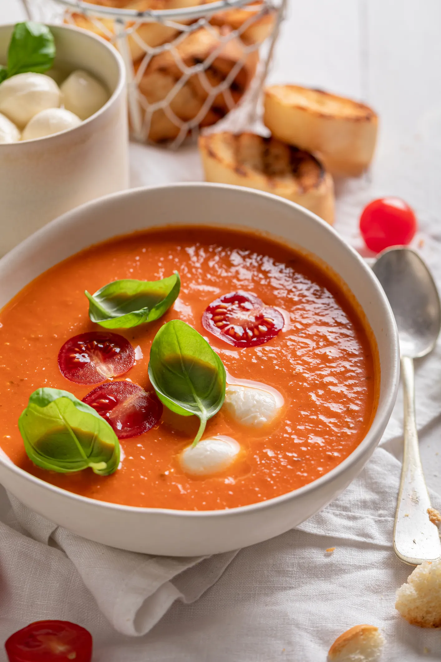 Beyaz bir masa örtüsü üzerinde, taze fesleğen yaprakları, dilimlenmiş çeri domatesler ve küçük mozzarella toplarıyla süslenmiş, kırmızımsı turuncu renkli bir kase Endülüs Gazpacho çorbası, yanında kızarmış ekmek dilimleri ve kasenin hemen sağında parlak bir kaşık görülüyor.