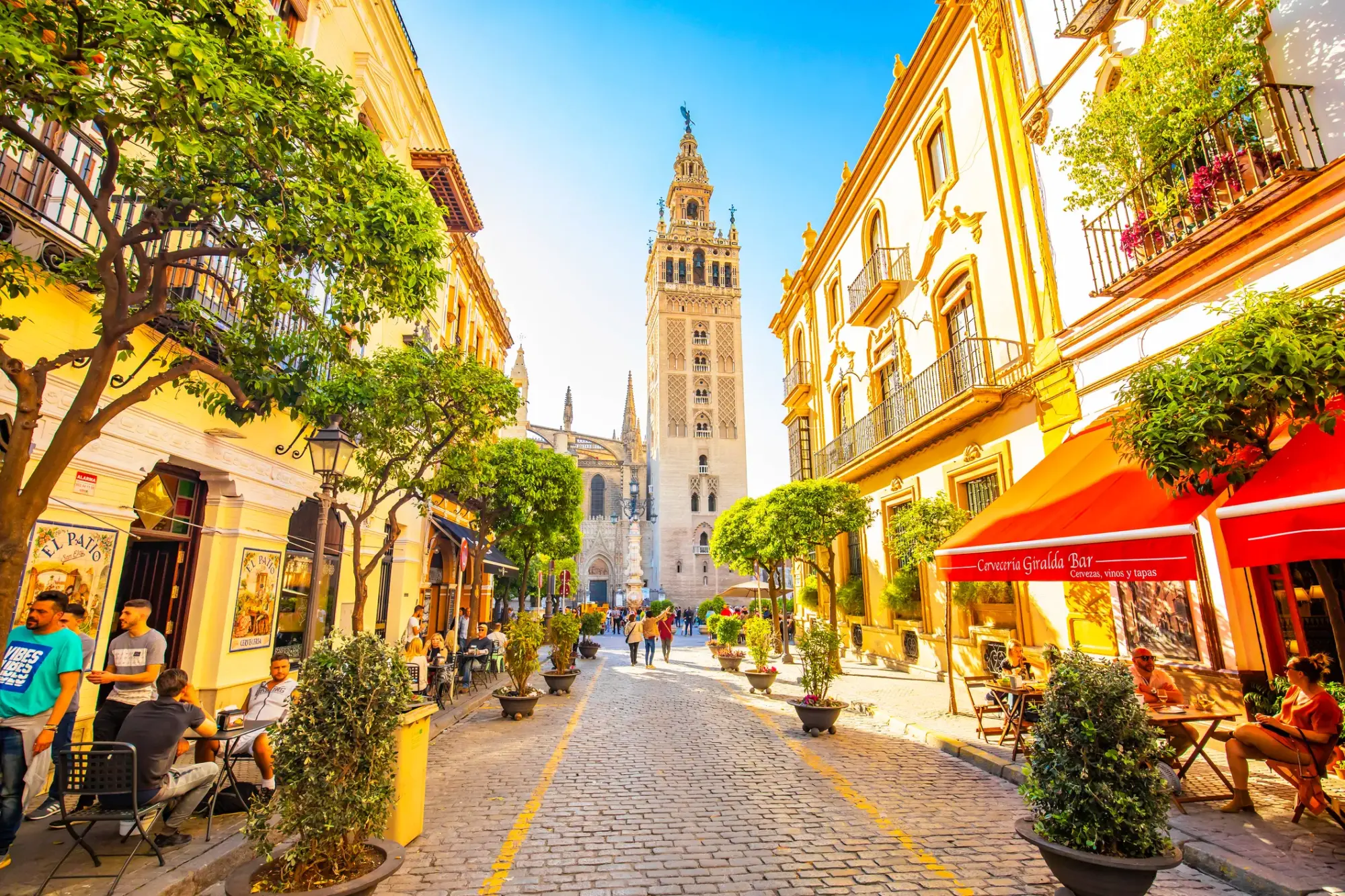 Güneşli bir günde, Sevilla'nın karakteristik dar arnavut kaldırımlı sokağında, dışarıya taşan masalarda oturan ve yürüyen insanların olduğu, ağaçlar ve çiçeklerle süslü binaların arasında yükselen Giralda Kulesi, şehri keşfedenler için sıcak bir atmosfer sunuyor.