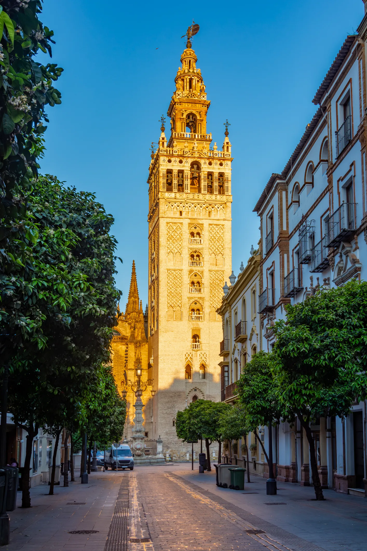 Altın rengi güneş ışıklarıyla aydınlanan Sevilla Katedrali'nin çan kulesi Giralda, yemyeşil ağaçların ve geleneksel İspanyol binalarının arasından göğe yükseliyor, ziyaretçilere unutulmaz bir seyahat rehberi deneyimi sunuyor.