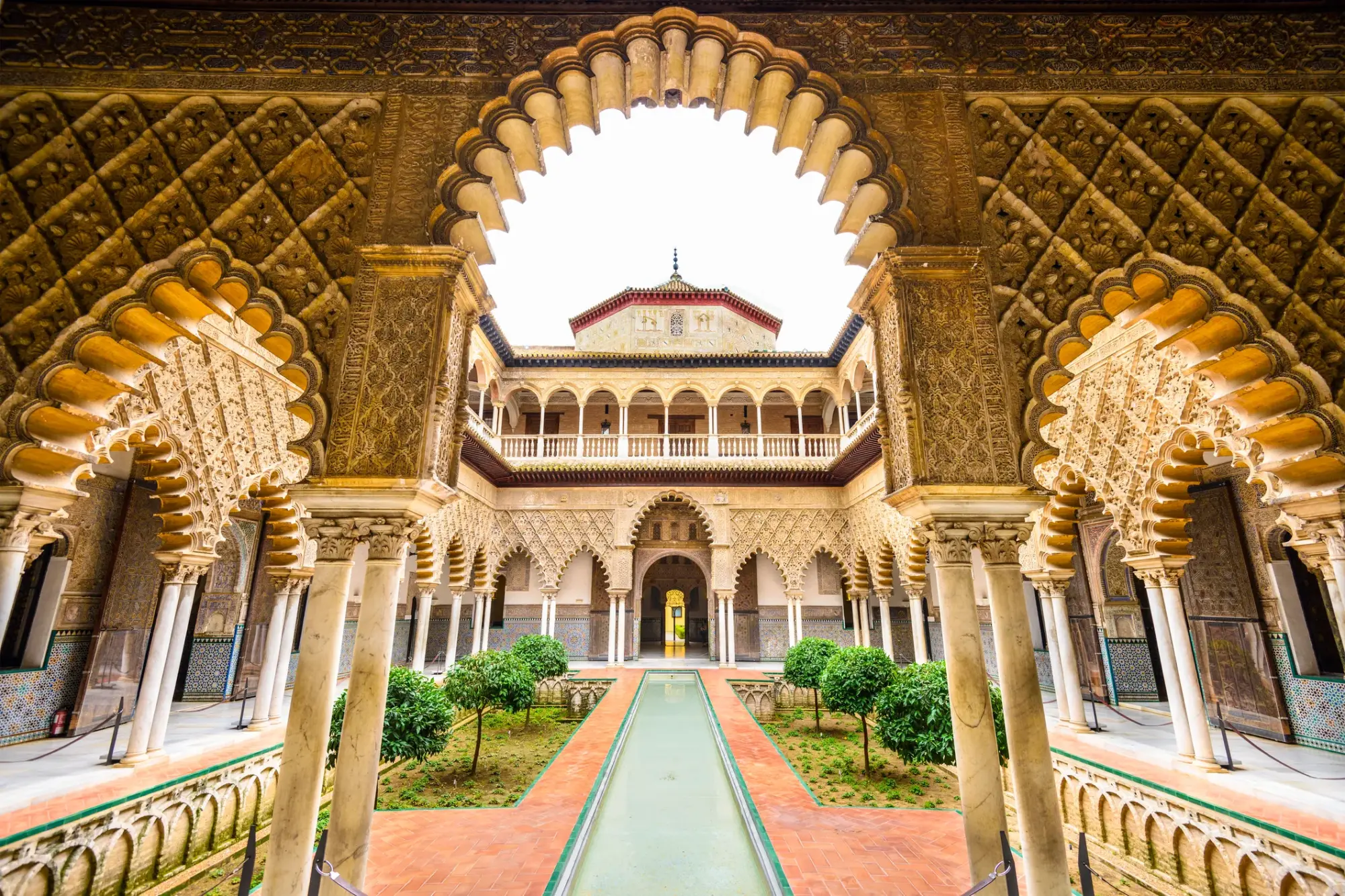 Sevilla'nın tarihi dokusunu yansıtan bu geniş avlu fotoğrafında, geleneksel İslam mimarisiyle bezeli oymalı kemerler, ortasında uzun bir su kanalı ve bakımlı bahçeler görünüyor, arka planda ise daha fazla katlı bir yapı yükseliyor.