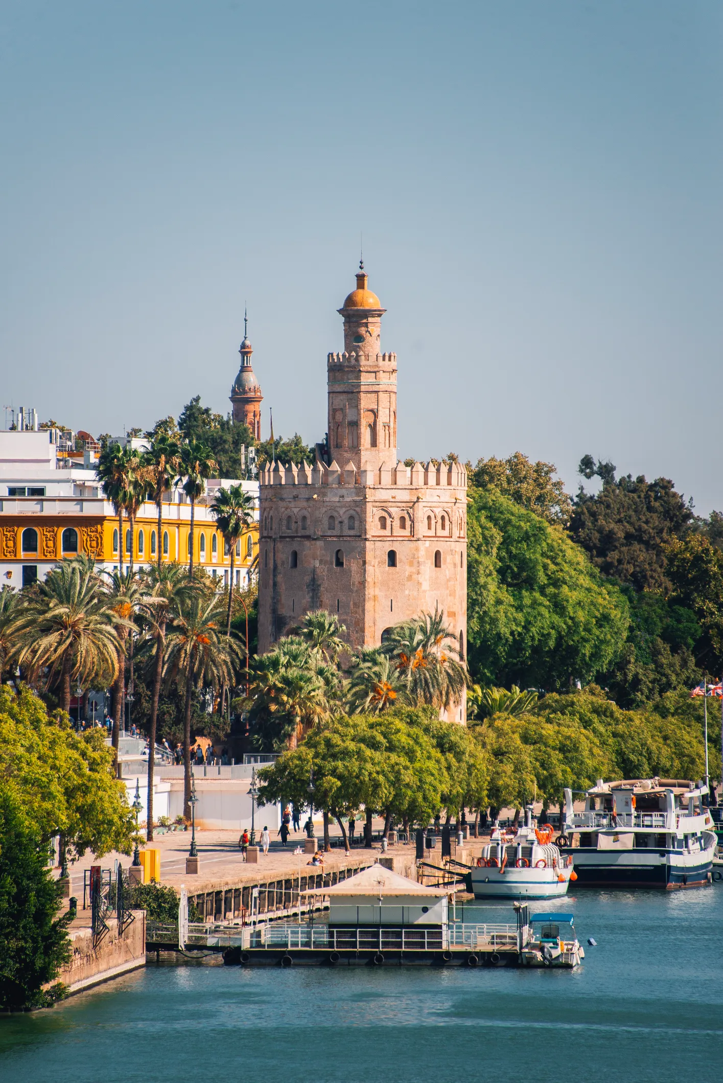 Sevilla'nın nehir kenarındaki popüler cazibe merkezlerinden biri olan altın kubbeli tarihi Torre del Oro kulesi, yemyeşil palmiye ağaçları, hareketli bir gezinti yolu ve demirli teknelerle dolu masmavi Guadalquivir Nehri üzerinde parlayan güneşli bir manzarada yer alıyor.