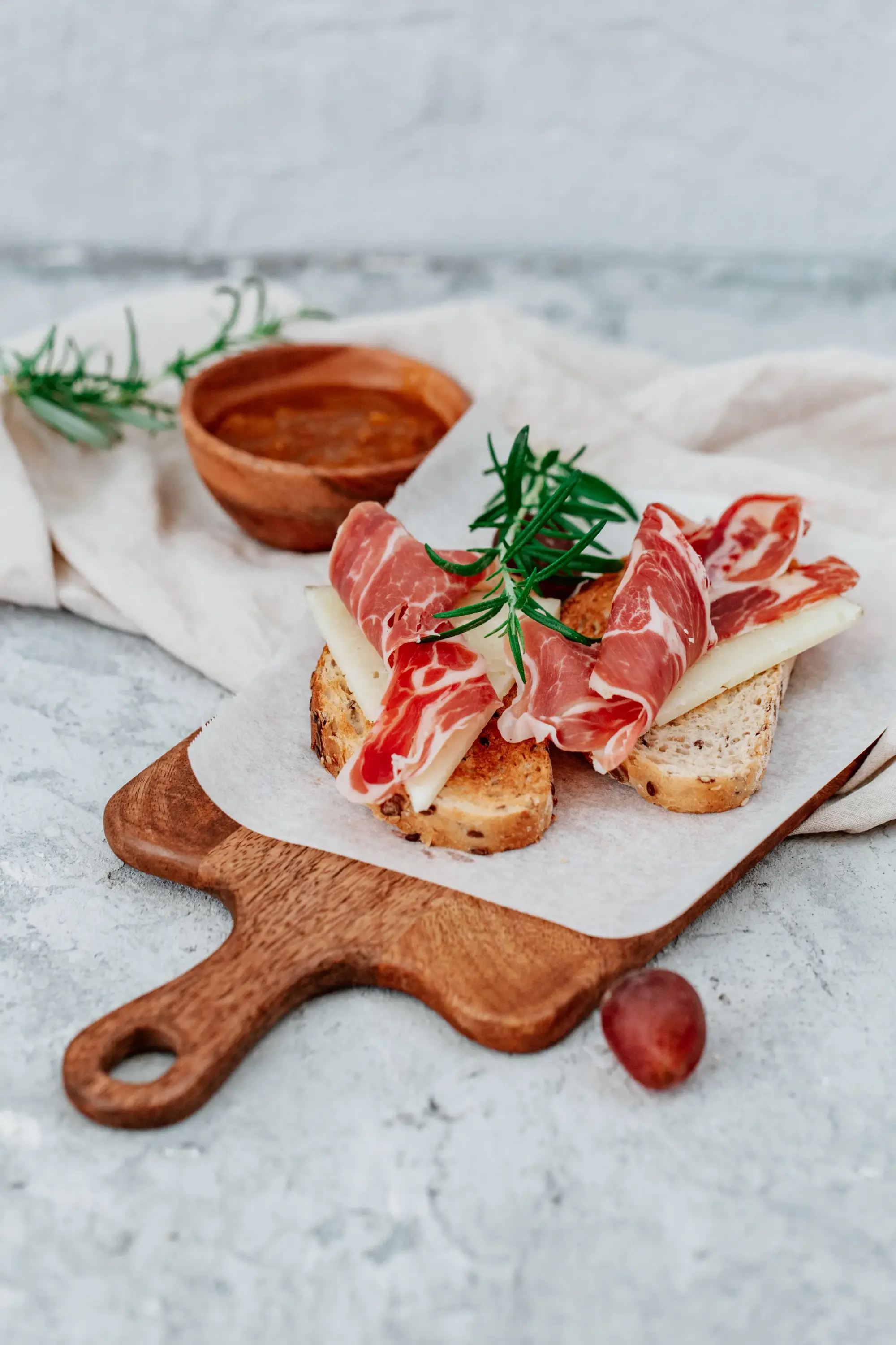 Ahşap servis tahtası üzerinde, kızarmış ekmek dilimlerinin üzerinde dilimlenmiş Manchego peyniri ve İspanyol jambonu, taze biberiye ile süslenmiş, yanında bir kırmızı üzüm ve arka planda sos kasesi ile keten örtüyle tamamlanan iştah açıcı bir Sevilla tapas tabağı.