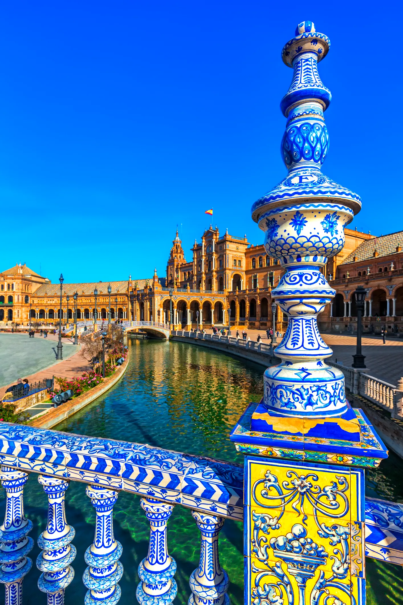 Sevilla'ya nasıl gidilir makalesine eşlik eden bu fotoğraf, ön planda mavi beyaz detaylı çini sütunu ve korkuluğuyla, arkasında berrak su kanalı ve güneşli havada parlayan muhteşem İspanya Meydanı mimarisini gösteriyor.