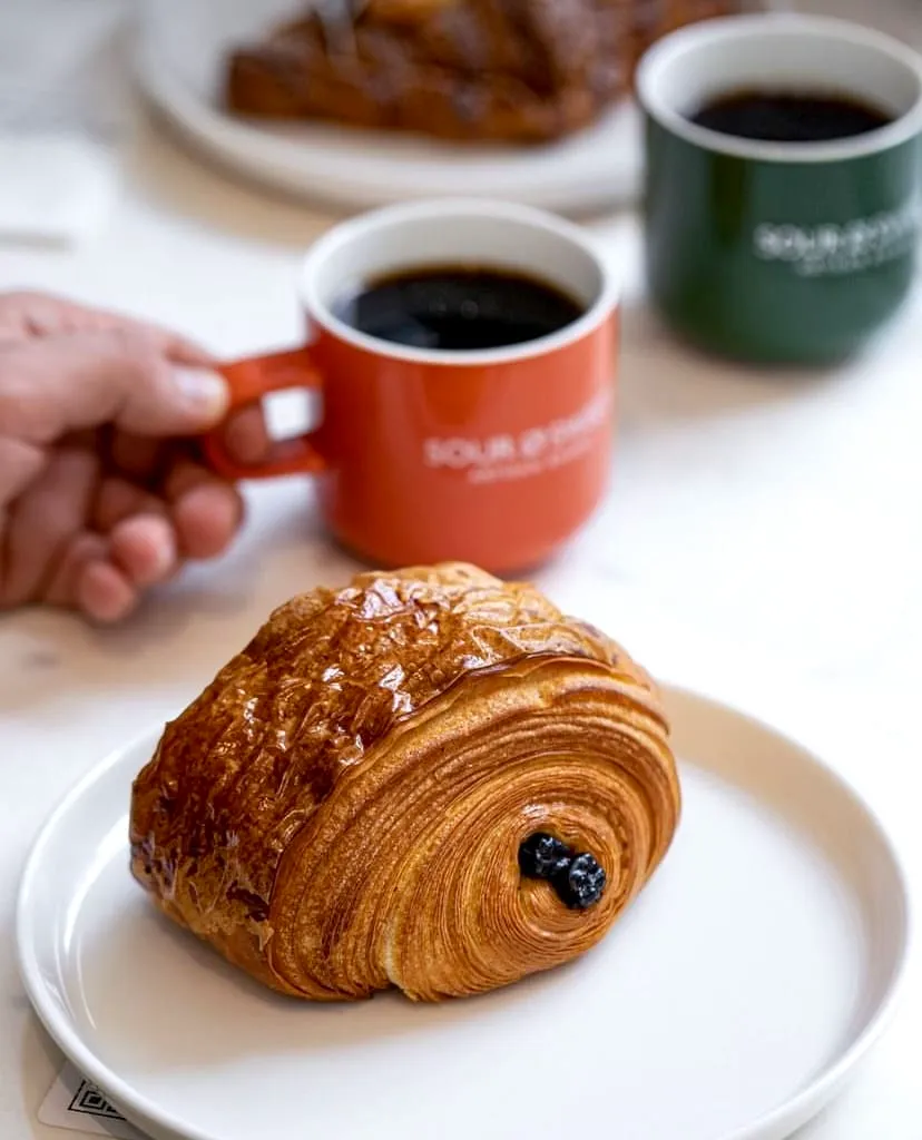 Beyaz bir tabağın üzerinde sunulan, kat kat dokusu ve parlak yüzeyiyle öne çıkan, orta kısmında koyu renkli meyveler barındıran lezzetli bir artisan hamur işi, Sour & Sweet Artisan Bakery logolu turuncu bir kupadaki sıcak kahveyle eşlik ederken, arka planda yeşil bir kupa ve hafifçe bulanık başka bir fırın ürünü de fark ediliyor.