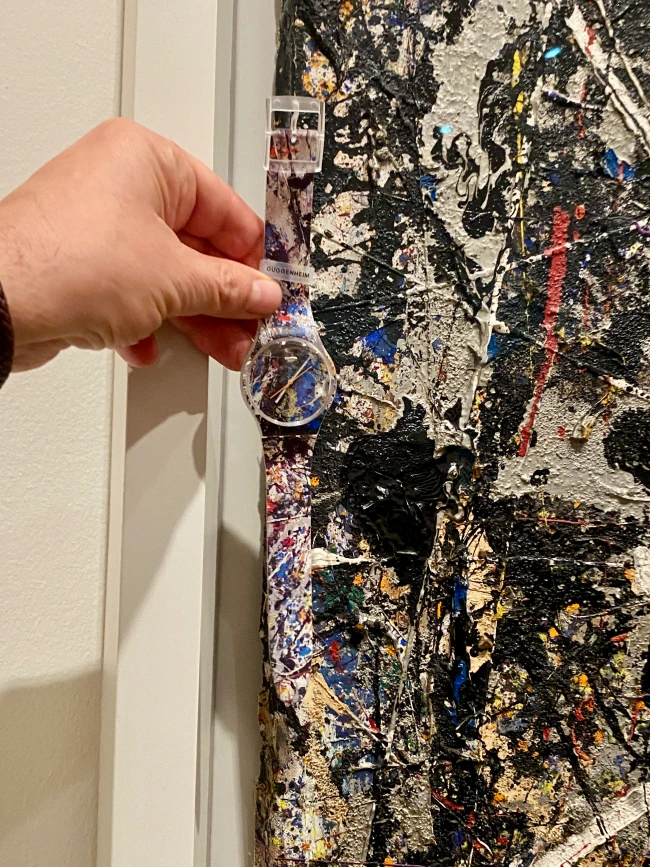 Bir el, Jackson Pollock'un resim stilinden esinlenilmiş, yoğun dokulu, boya sıçramalarıyla dolu soyut bir tablonun önünde Swatch Jackson Pollock Alchemy saatini tutuyor; saatin kordonu ve kadranı da tablonun renkli ve dinamik desenlerini taşıyor, kordonda küçük bir GUGGENHEIM etiketi görülüyor.