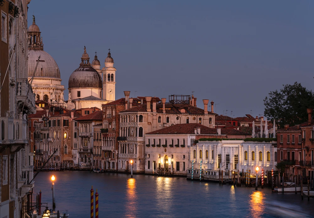 Venedik'in Büyük Kanalı boyunca uzanan, akşam alacakaranlığında ışıklarla aydınlanmış tarihi binaları, sol tarafta Santa Maria della Salute kubbesi ve sağda belirginleşen Peggy Guggenheim Koleksiyonu'nun cephesini gösteren büyüleyici bir şehir manzarası.