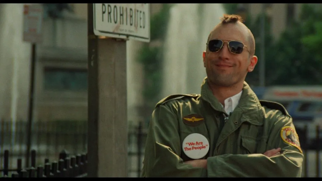 Taxi Driver filmindeki ikonik Travis Bickle karakterini canlandıran Robert De Niro, mohawk saç kesimi, güneş gözlüğü ve üzerinde We Are The People yazılı bir rozet bulunan haki askeri ceketiyle, kolları bağlı ve hafifçe gülümseyerek kalabalık bir şehir sokağında duruyor.
