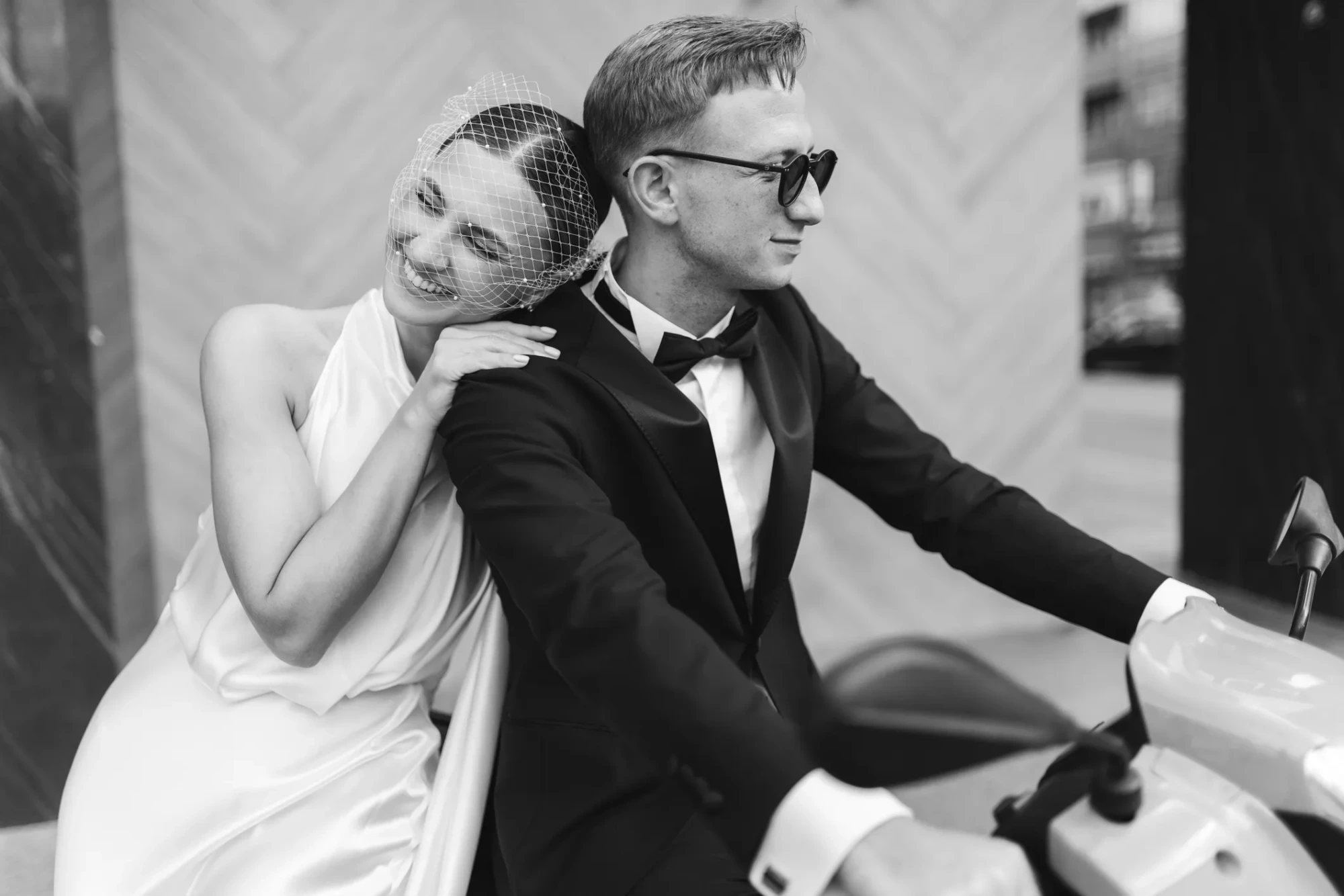 Siyah beyaz fotoğrafta, smokinli ve güneş gözlüklü bir adam scooter üzerinde otururken, neşeli bir gülümsemeyle duvaklı bir kadın omzuna yaslanmış, Terazi burcunun aşk hayatında yeni başlangıçları ve derin bağlılıkları işaret ediyor.