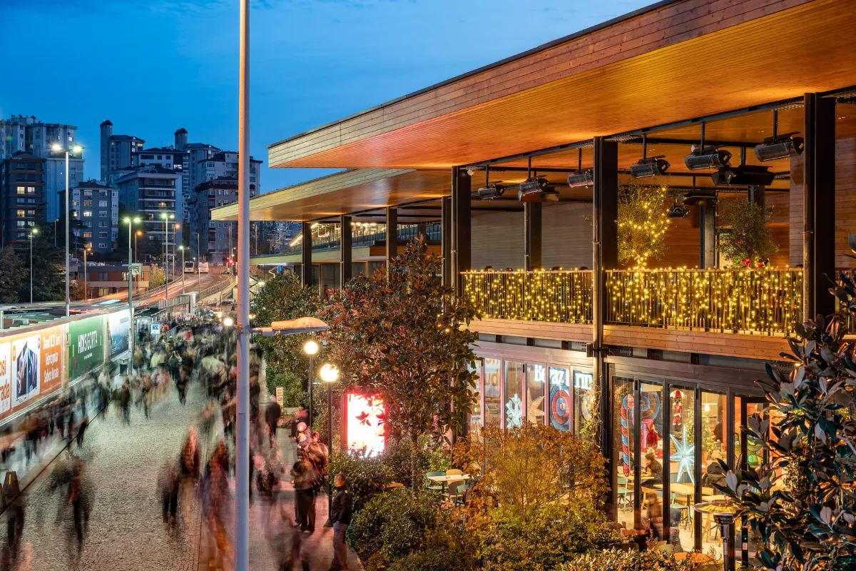 Akşamüstü ışıkları altında, Kadıköy'ün dinamik atmosferini yansıtan Terminal Kadıköy'de, hareketli bir cadde boyunca yürüyen insan kalabalığı ve altın sarısı ışıklarla süslenmiş modern balkonlar, buranın canlı bir buluşma noktası olduğunu gösteriyor.