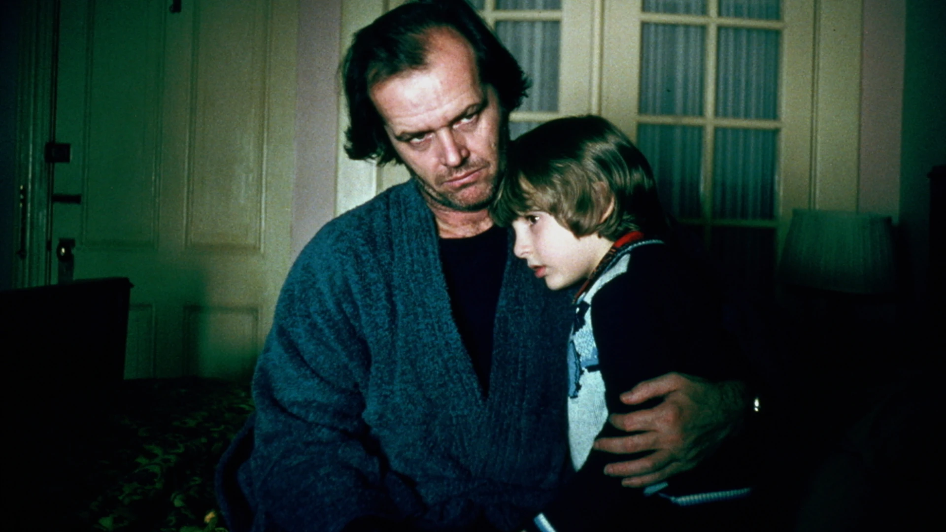 The Shining filminden bir sahne: Gergin ve endişeli bir ifadeyle doğrudan kameraya bakan, Jack Nicholson'ın canlandırdığı Jack Torrance, loş yeşilimsi ışıkla aydınlatılmış bir odada, ürkmüş görünen genç oğlu Danny'yi kollarında tutuyor. Danny'nin endişeli yüzü babasının omzuna dayanmış, kenara bakıyor.