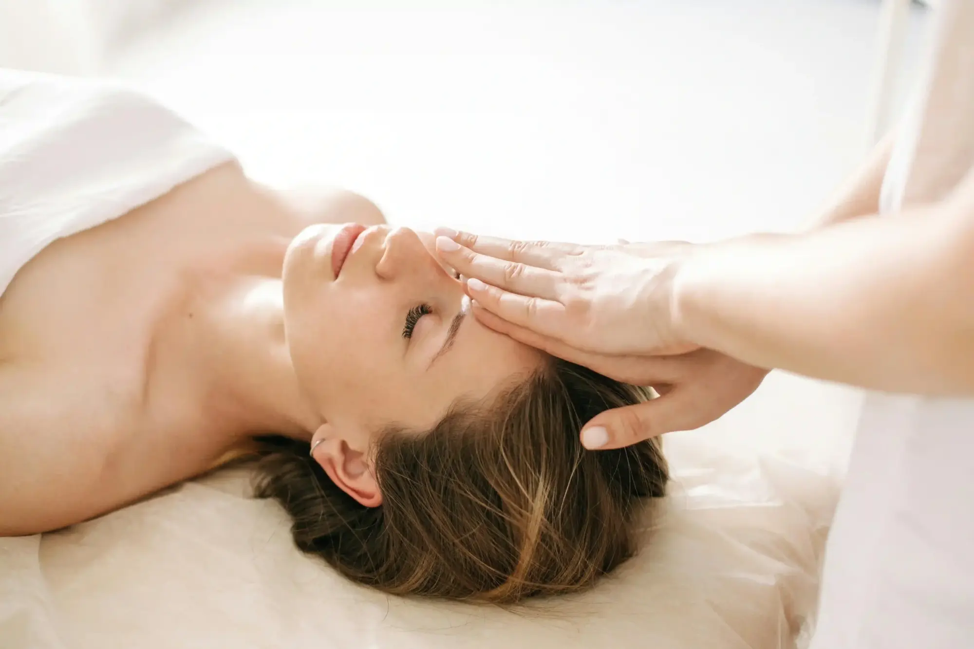Gözleri kapalı bir kadın yatakta uzanırken, bir uygulayıcının eli nazikçe alnına dokunarak bilinçaltı inanç dönüşümüne odaklanan bir Theta Healing seansında derin bir meditasyon ve rahatlama anını görselleştiriyor.