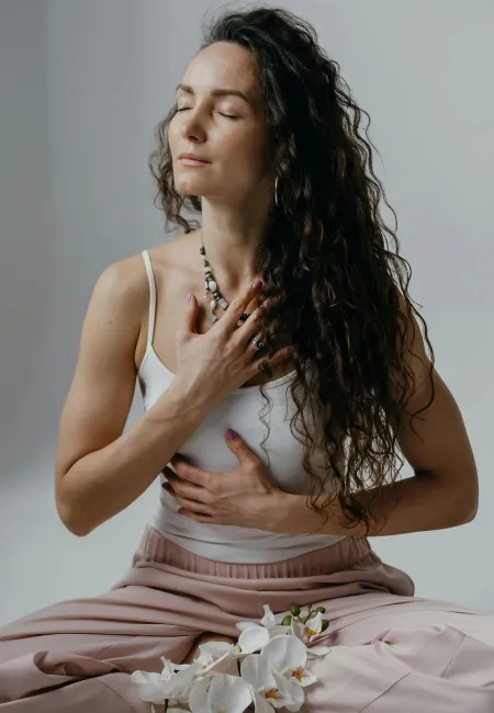 Uzun kıvırcık saçlı, beyaz bir atlet ve pembe pantolon giyen kadın, gözleri kapalı, elleri göğsünde meditasyon pozisyonunda bağdaş kurmuş olarak derin bir içsel odaklanma ve huzur içinde görünüyor; önündeki zarif beyaz orkidelerle birlikte bu görüntü, Theta Healing'in temelini oluşturan derin gevşeme ve bilinçaltı inanç çalışmasını doğal bir şekilde yansıtıyor.