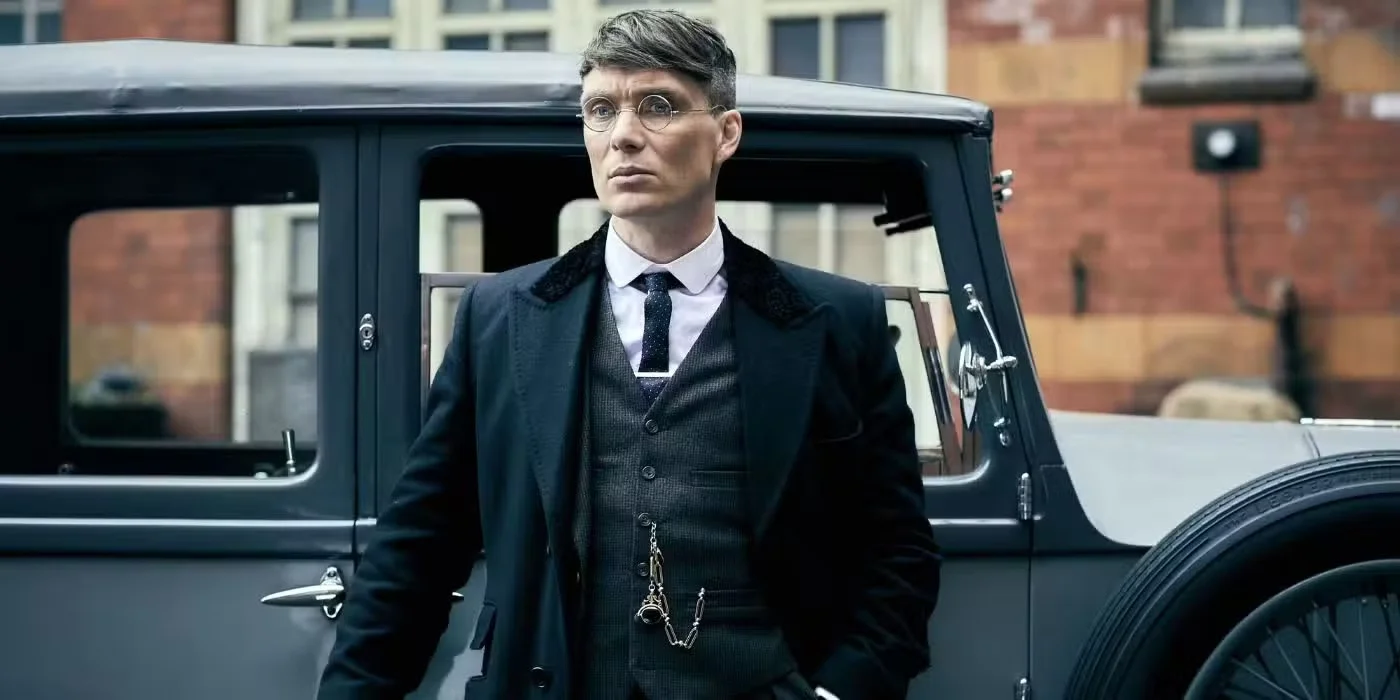 Peaky Blinders karakteri Tommy Shelby, dönemin modasına uygun üç parçalı takım elbisesi, uzun paltosu ve yuvarlak gözlükleriyle, arka planda tuğla binaların seçildiği bir sokağın kenarında park etmiş koyu renk lüks bir klasik otomobilin yanında ciddi bir ifadeyle duruyor.