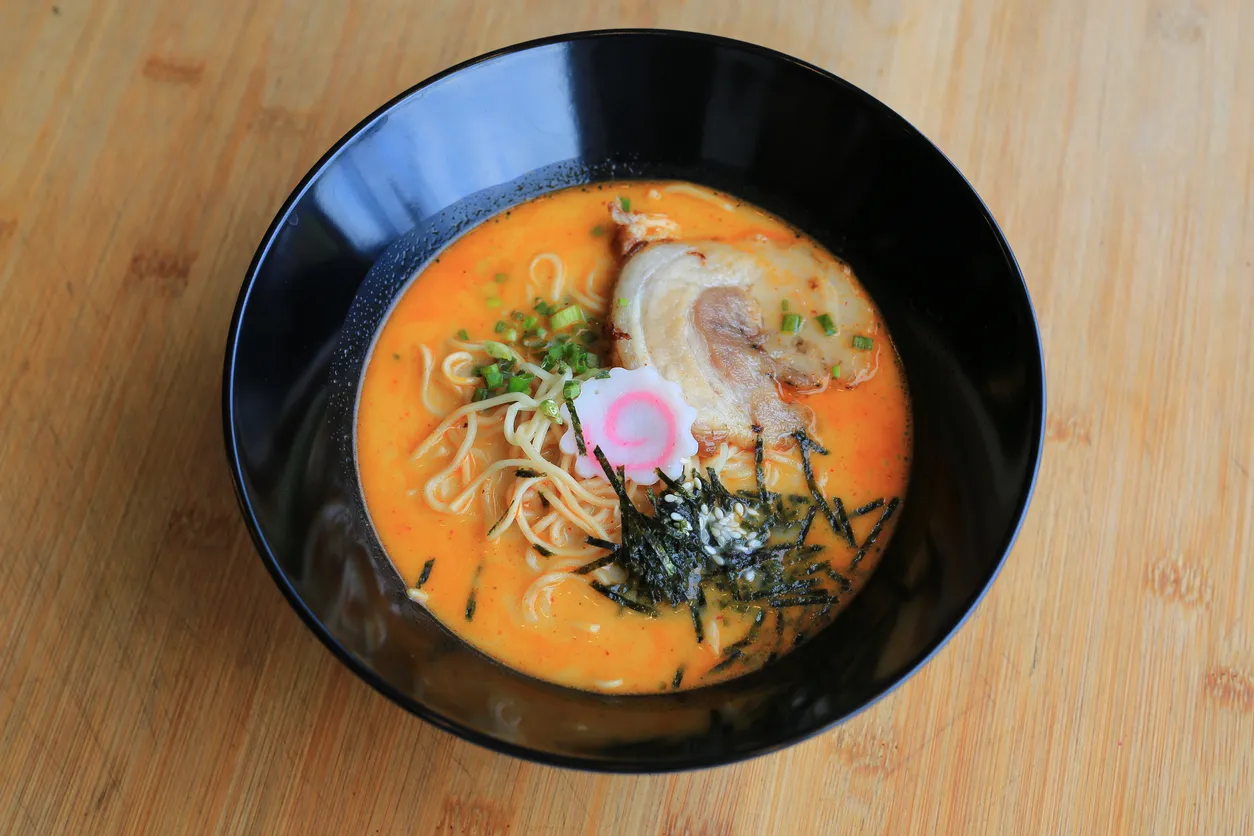 Oggusto'nun Ramen Nedir? Shoyu, Miso ve Daha Fazlası rehberindeki zengin umami kültürünü yansıtan siyah kase. Ahşap masada duran bu iştah açıcı sunumda; kremsi turuncu çorba, taze erişteler, et dilimi, pembe sarmallı narutomaki, nori şeritleri, taze soğan ve susam taneleri bir araya geliyor.