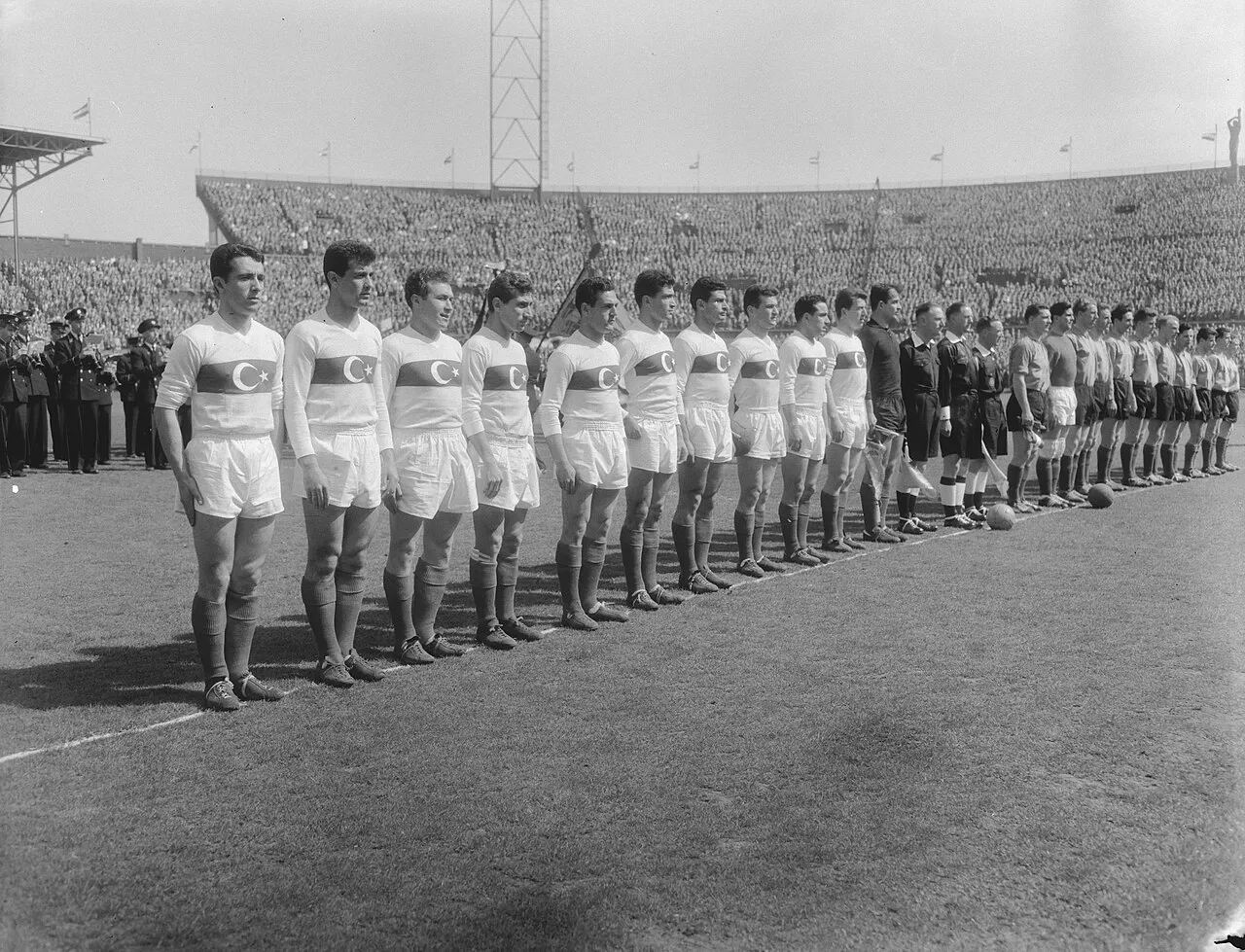 1958 Türkiye-Hollanda milli maçı öncesi, beyaz formalı Türkiye milli futbol takımının ve rakip Hollanda takımının sahada sıraya dizilişini, aralarındaki hakemleri ve arka planda tribünleri dolduran kalabalık seyircileri gösteren siyah beyaz bir fotoğraf.