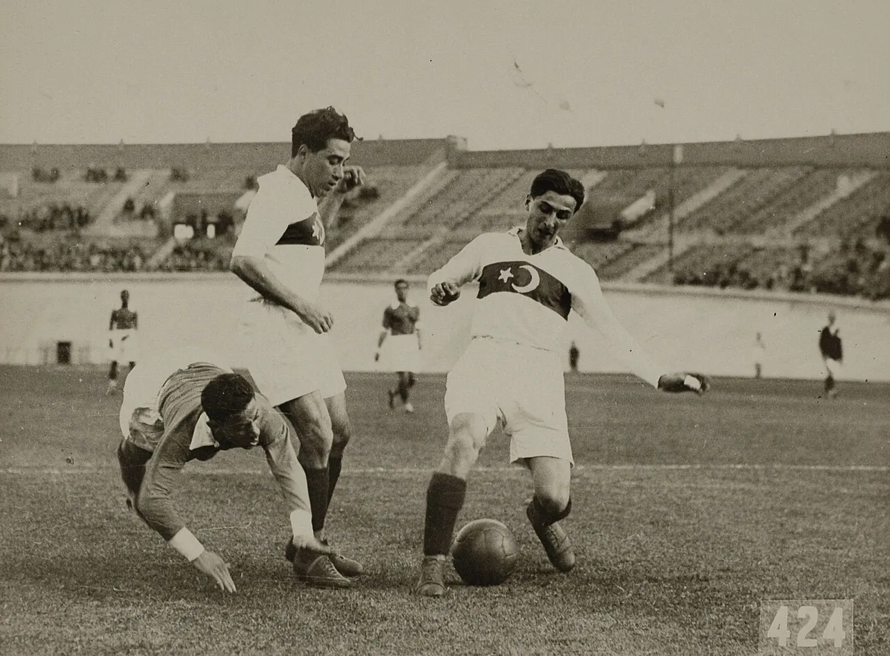 Siyah beyaz bu tarihi fotoğraf, 1928 Olimpiyat Oyunları'ndaki Türkiye-Mısır futbol maçından heyecanlı bir anı ölümsüzleştiriyor; beyaz ay yıldızlı formasıyla top süren Türk oyuncunun önünde eğilerek topa müdahale etmeye çalışan Mısırlı bir rakip ve arkasında bir takım arkadaşı, seyircilerin de olduğu stadyumda çim sahada mücadele ediyor.