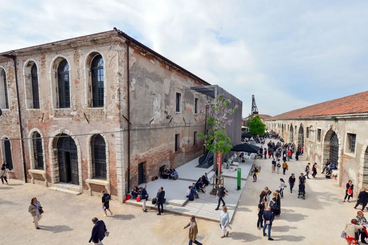 Venedik Bienali Türkiye Pavyonu'nun yer aldığı, eski tuğla ve sıvalı binaların çevrelediği geniş bir açık alanda, mor salkımlı bir ağacın ve modern ahşap yapıların etrafında toplanmış kalabalık ziyaretçiler yürüyüp otururken, uzakta endüstriyel bir vinç görülüyor.