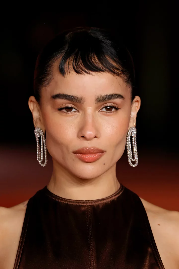 Zoe Kravitz, açık kahverengi dudakları, belirgin kaşları ve alnının ortasına kadar inen kısa, dalgalı mikro kakül saç modeliyle doğrudan kameraya bakarken, parıldayan oval uzun küpeleri ve kahverengi bir üst giyiyor.