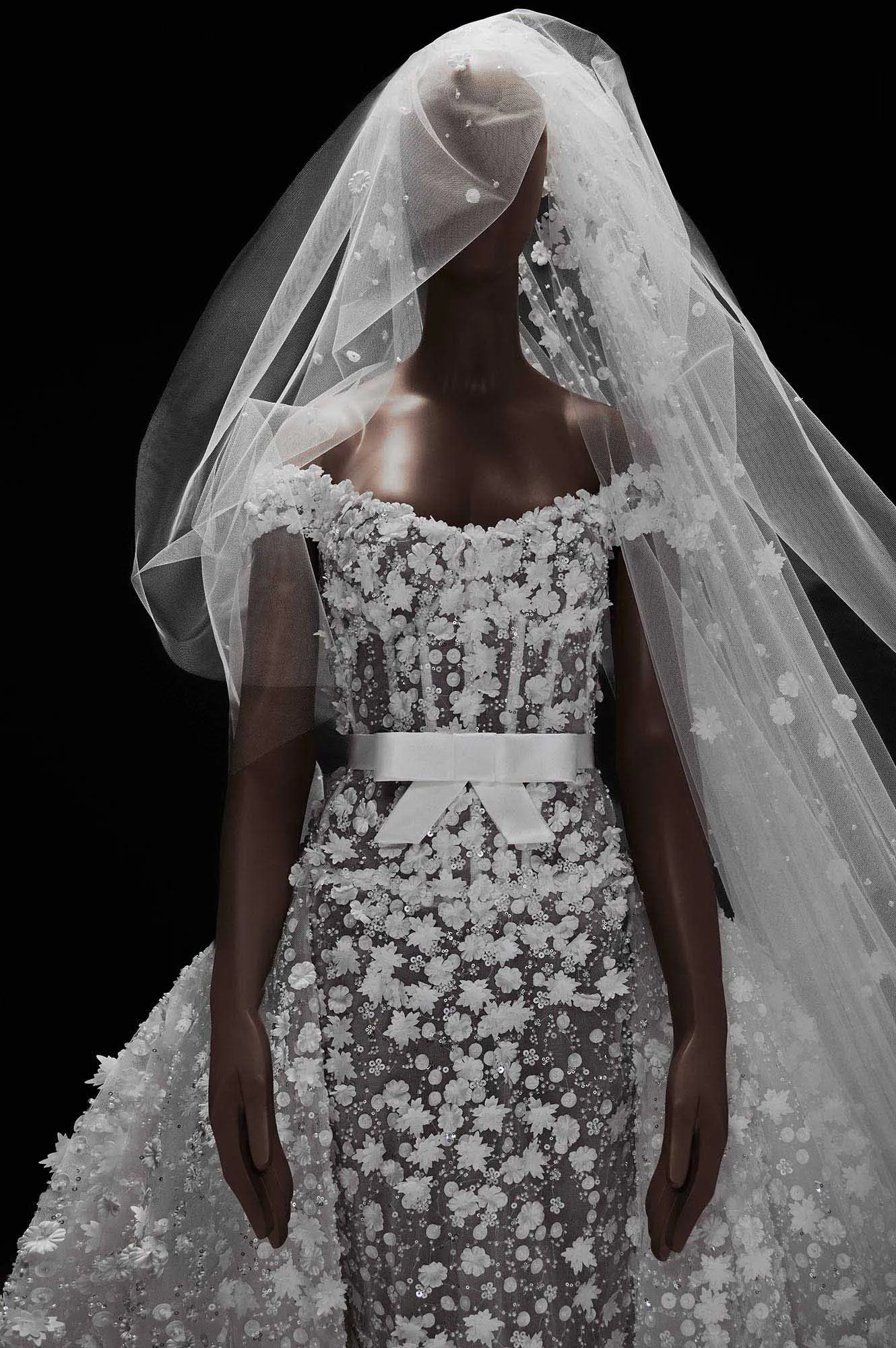Zuhair Murad'ın couture işçiliğiyle hazırlanan koleksiyonundan, zarif üç boyutlu çiçek aplikeleri ve payetlerle bezenmiş transparan dokulu, omuzları açıkta bırakan silueti beyaz bir kuşakla vurgulanan ve uzun bir duvağın eşlik ettiği, romantizmi modern bir siluetle buluşturan görkemli bir gelinlik.